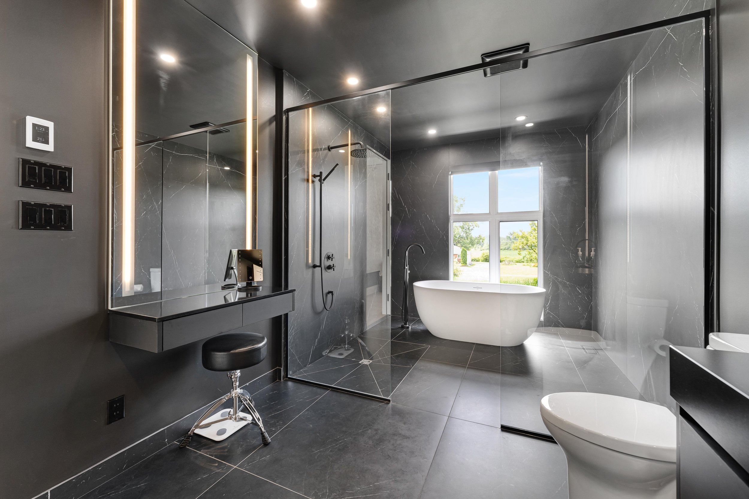 Modern bathroom with black marble design, featuring a glass walk-in shower, freestanding white bathtub, wall-mounted vanity with mirror, and black stool; large window with outdoor view.