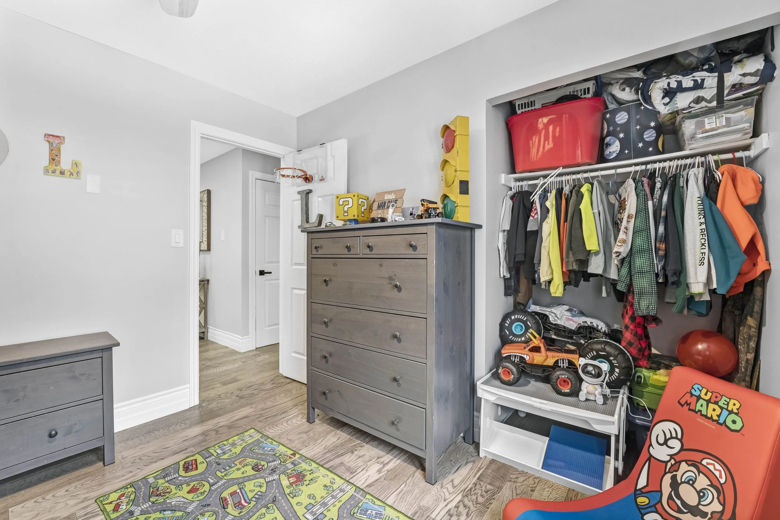 photographie immobiliere outaouis  Child's bedroom with gray furniture, toys, and clothing. Includes a dresser, a small table, a closet with a rod of hanging clothes, and toy vehicles. The floor has a colorful carpet featuring a road map design.