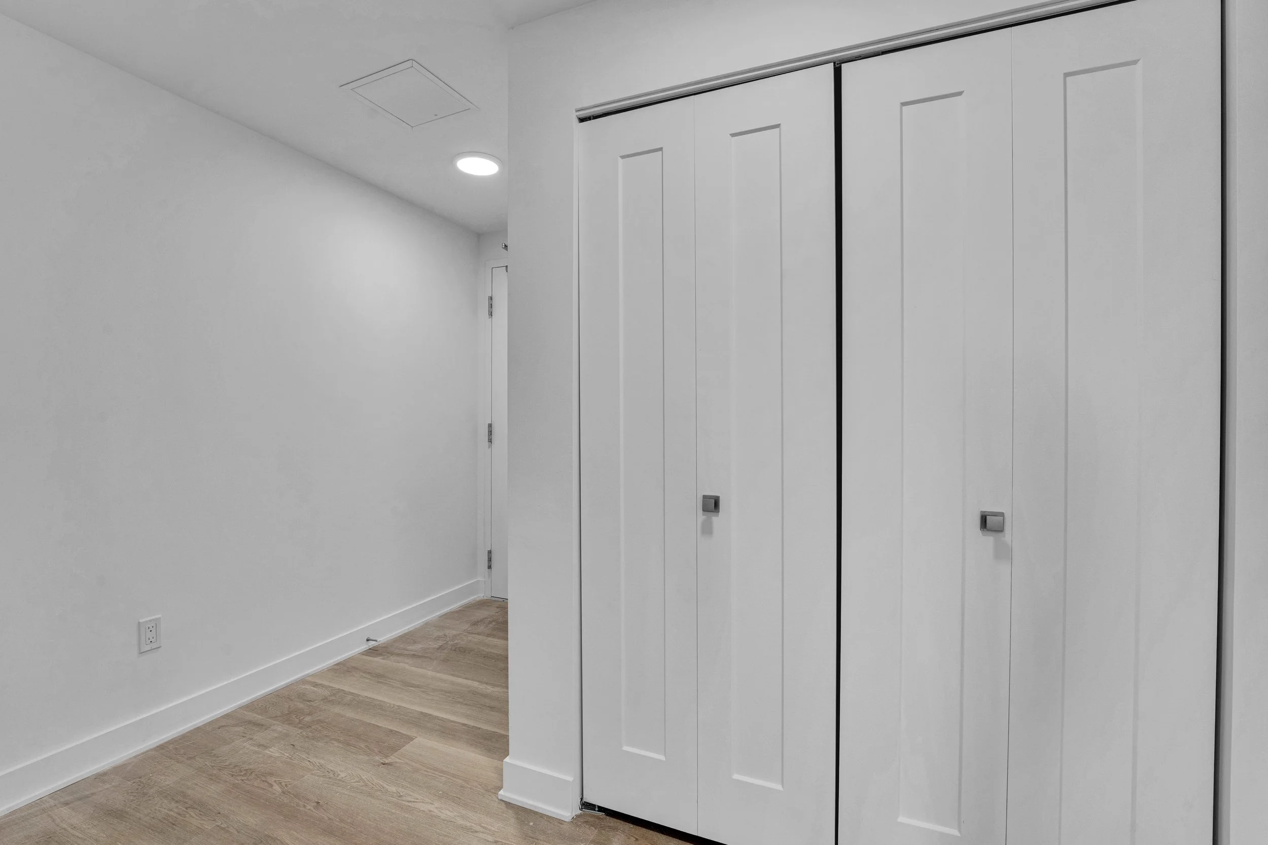 Ottawa Real Estate Videographer NVISION 3D  A minimalist room with white walls, wood flooring, a closed white door, and white closet doors with silver handles. There's a ceiling vent and a round ceiling light.