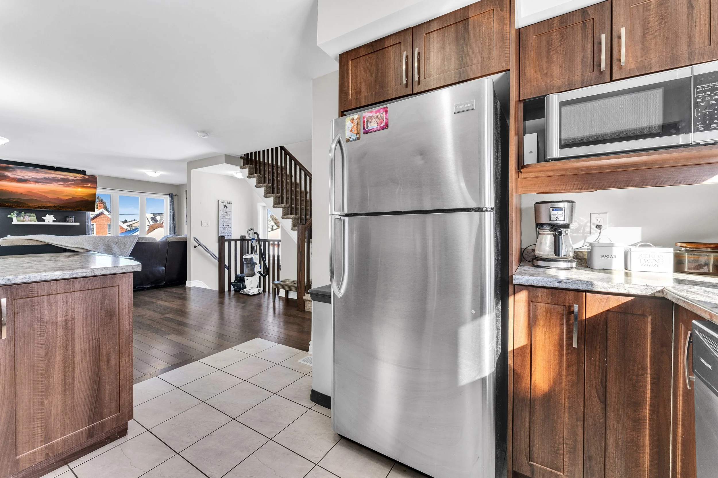 NV 3D Real Estate Media Ottawa  Stainless steel refrigerator in a kitchen with wooden cabinets, microwave, coffee maker, and various small appliances on the counter. Part of the living room with a TV and sofa is visible in the background.