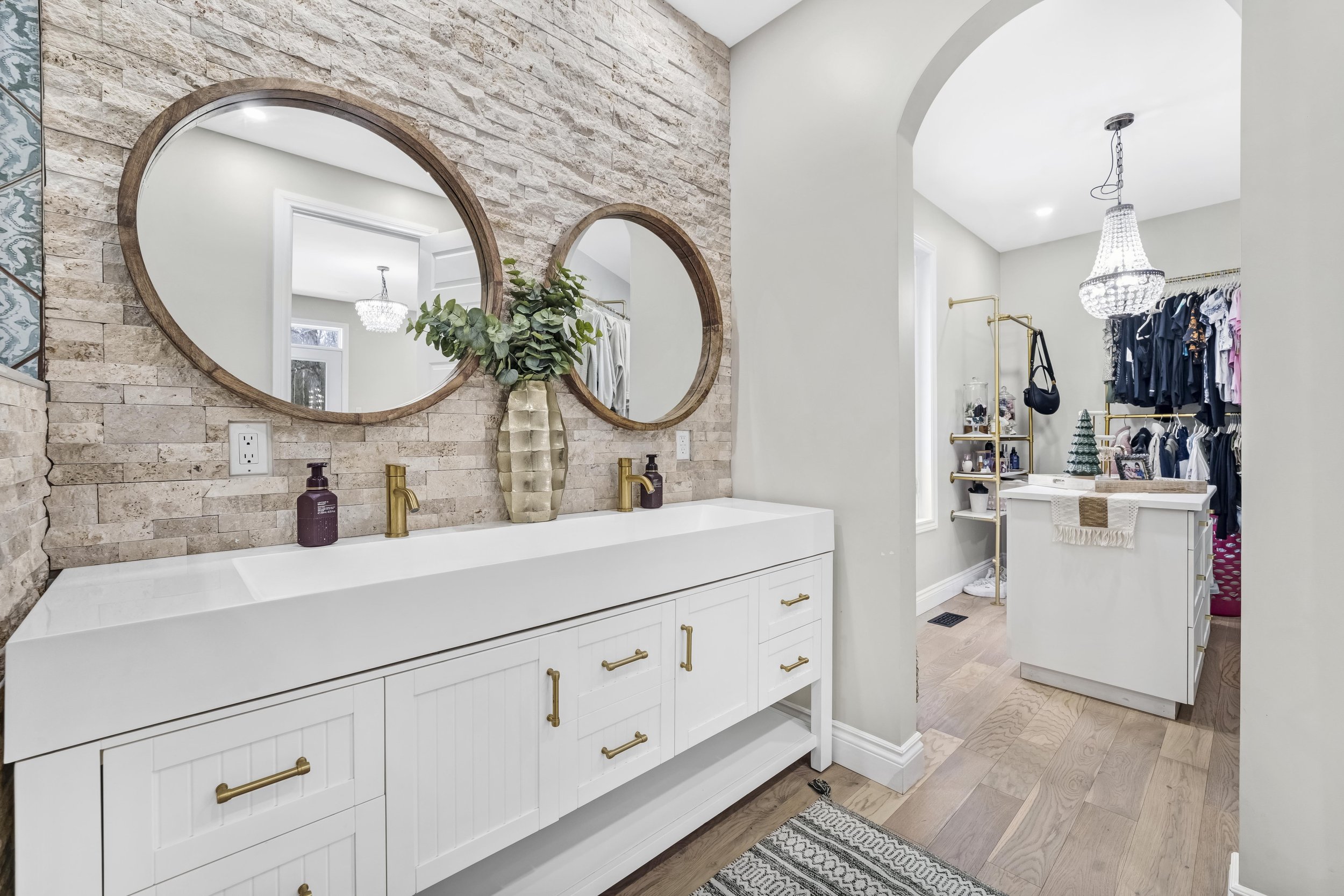 Bathroom with a double vanity, two round mirrors with wooden frames, a beige brick wall behind the vanity, a decorative plant in a gold vase, and a dual sink with gold fixtures. Adjacent to the bathroom is a walk-in closet with hanging clothes, a cha