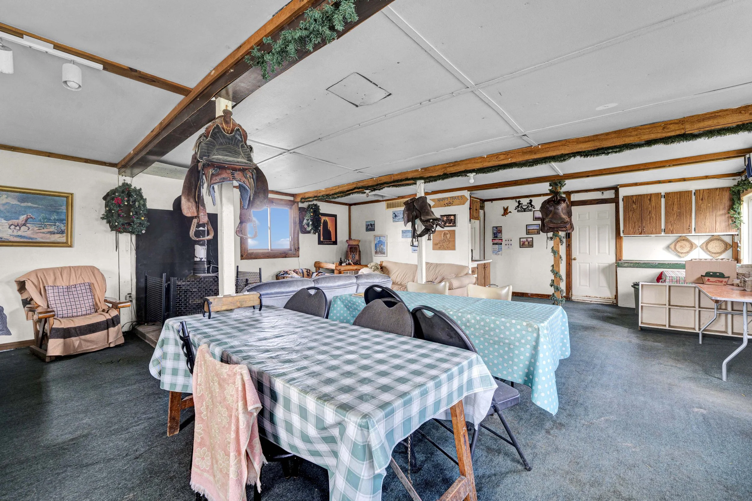 The image shows the interior of a rustic-style dining area with tables covered in green checkered tablecloths. There are various chairs around the tables, and the room's walls are decorated with framed art, some of which feature horses and scenic lan