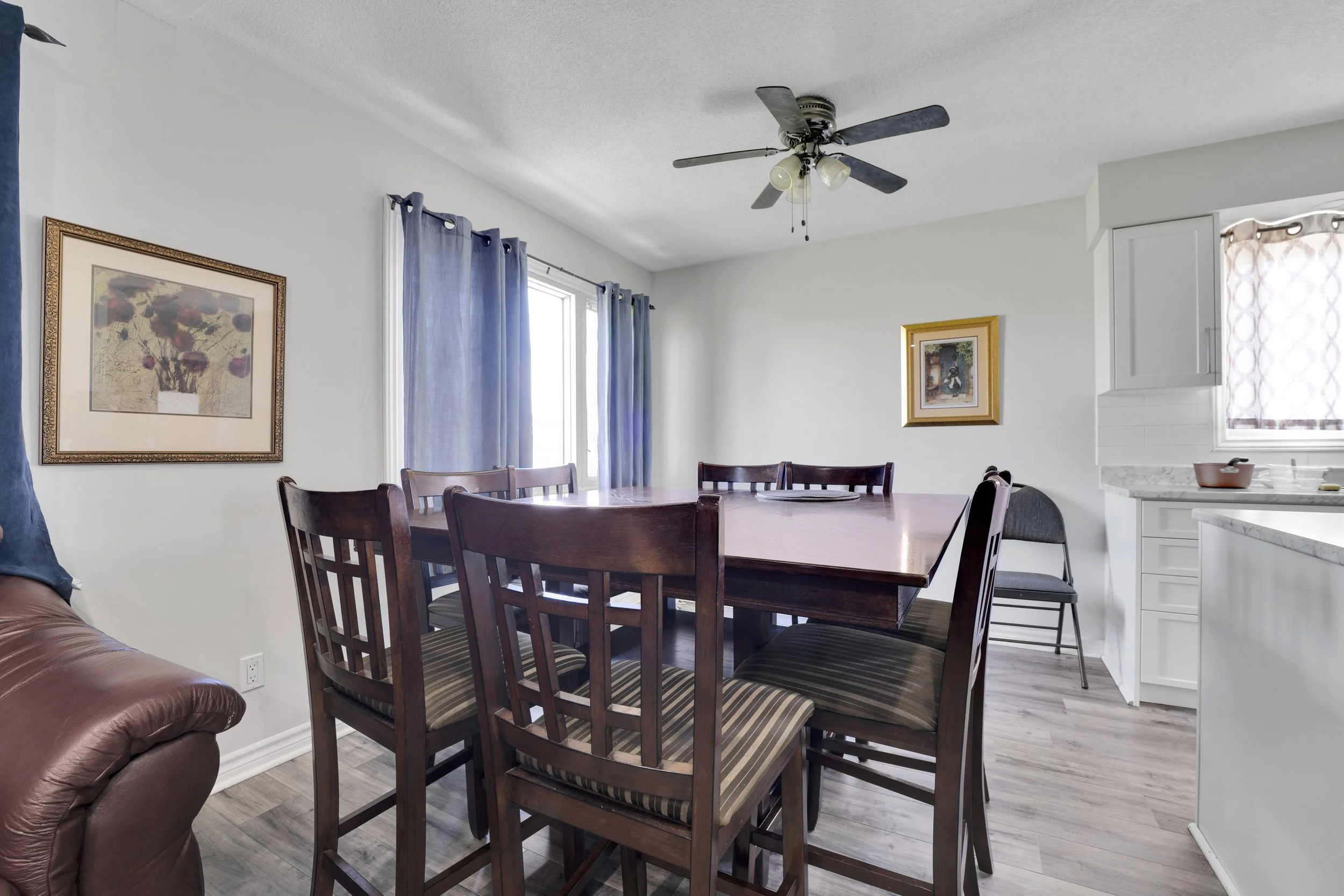 Ottawa Real Estate MInterior of a dining room with a wooden table surrounded by matching chairs, blue curtains on windows, framed artwork on white walls, a ceiling fan with lights, and a glimpse of the kitchen with white cabinetry and a small window.
