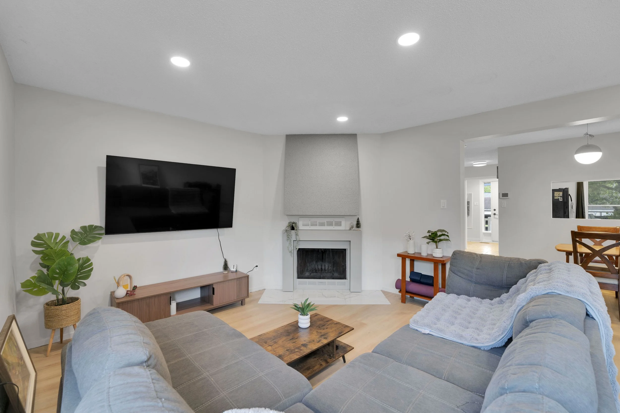 Ottawa Real Estate Video Services  Living room with gray sofa, wooden coffee table, potted plants, wall-mounted TV, fireplace, and dining area with round table and chairs, bright and airy with natural light.
