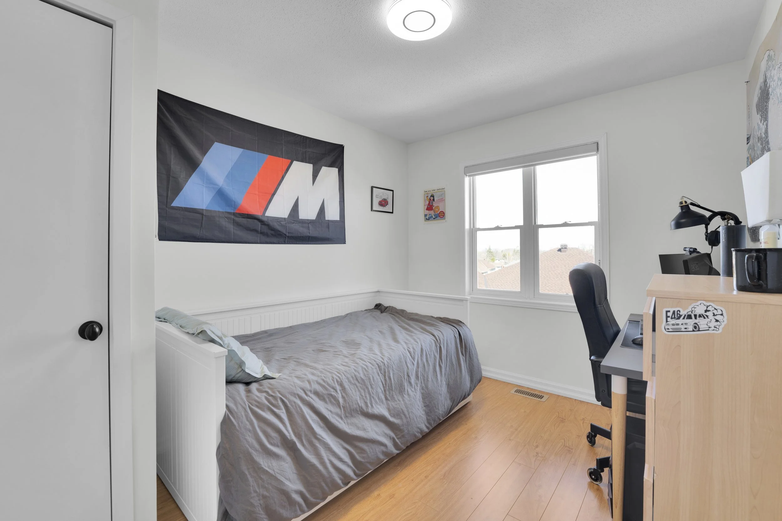 NVISION3D.ca Real Estate Media Ottawa  A bedroom with white walls, a window, a bed with gray bedding, a black chair, a wooden desk with a computer, a lamp, and a flag with the BMW M logo hanging on the wall.