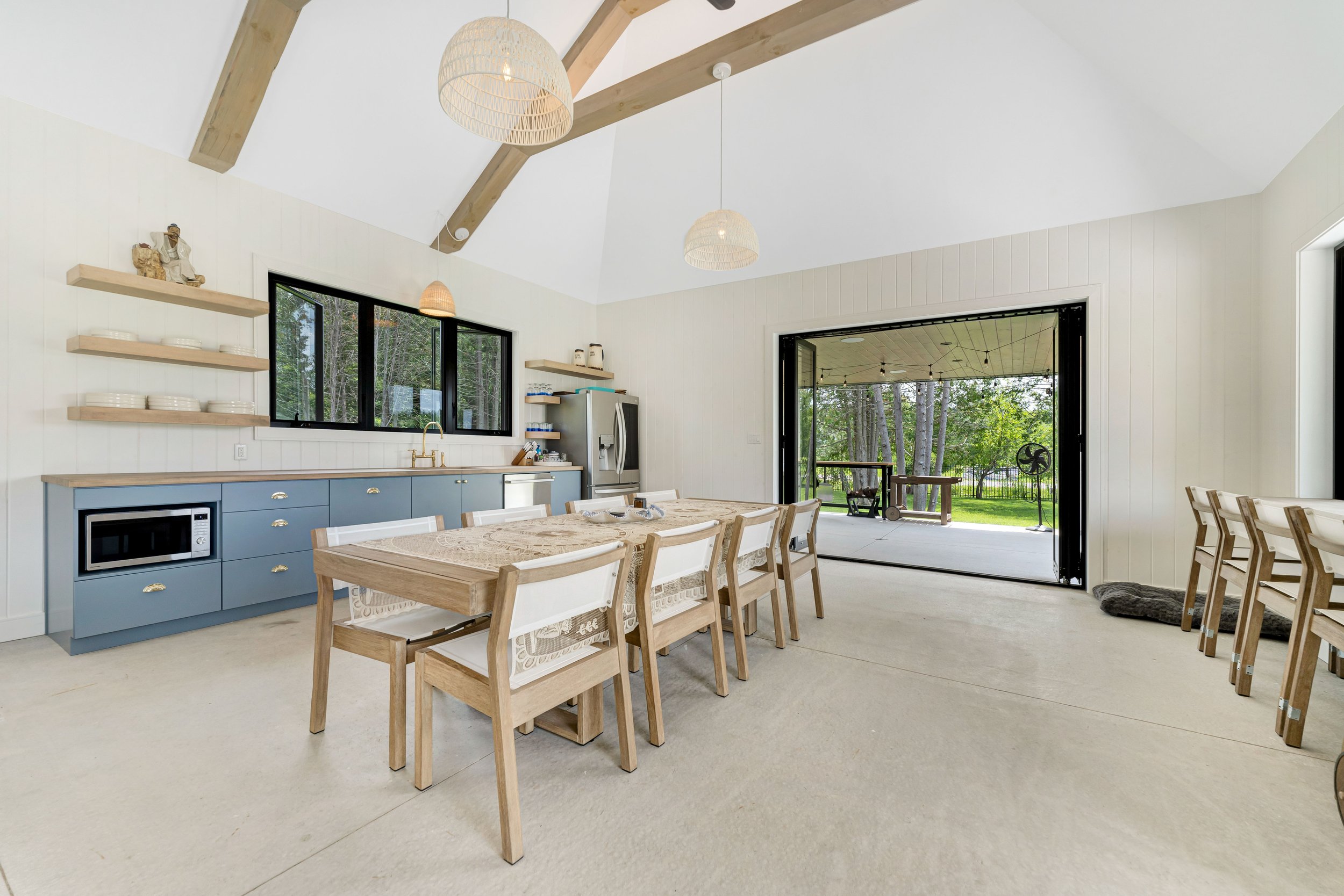 Modern dining area with wooden dining table, six chairs, blue cabinets, and open shelves. Large windows and doors open to a porch with views of trees. Pendant lights hang from the ceiling, and a microwave is integrated into the cabinets.
