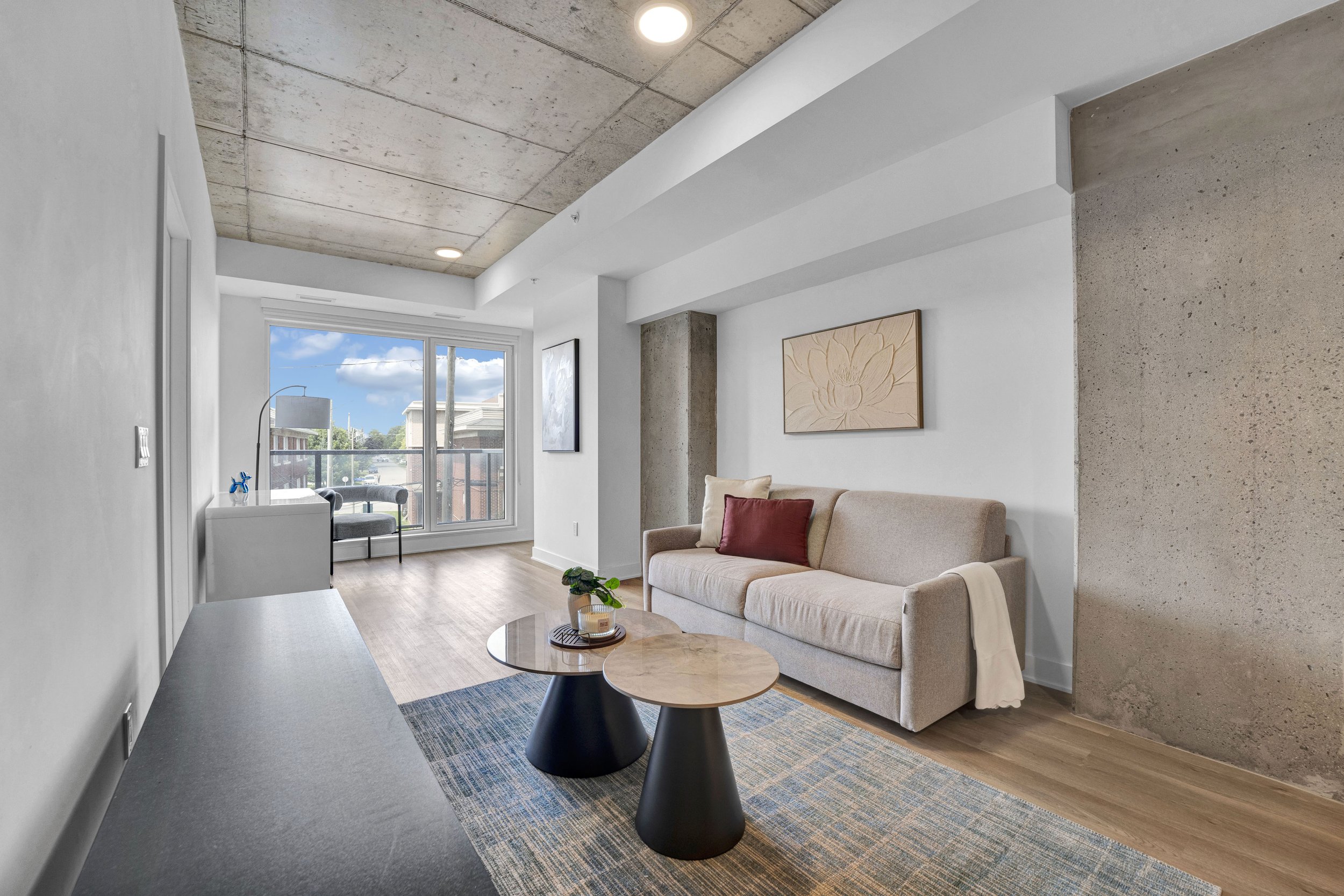 Ottawa Real Estate NVISION 3D  Modern living room with a beige couch, red and cream cushions, a coffee table with candles, artwork on white walls, large window, concrete and white ceiling, and hardwood floor.