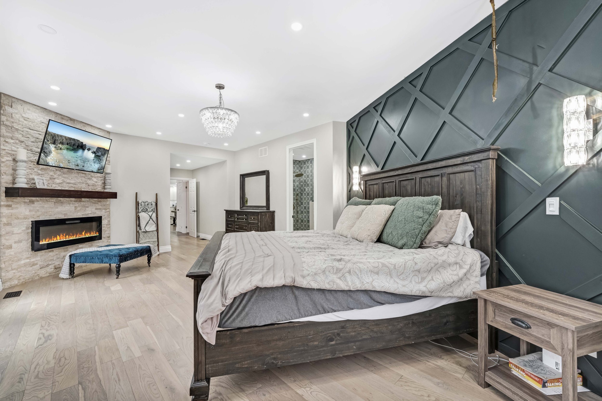 photographie immobiliere outaouis  Modern bedroom with a large wooden bed, dark blue accent wall, and fireplace with a TV above. Light wood flooring and a chandelier hanging from the ceiling.