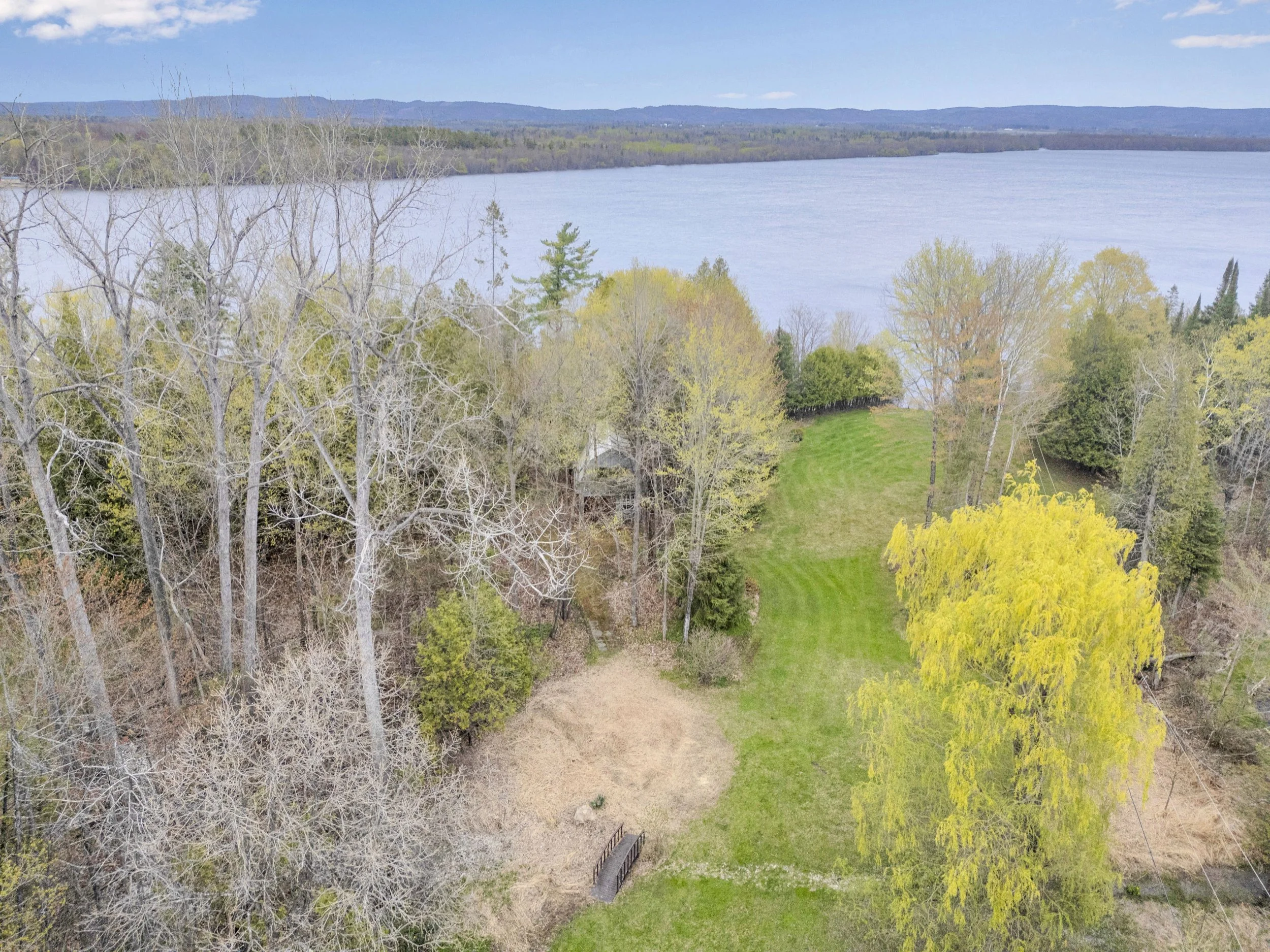 Ottawa Real Estate Media Services  Aerial view of a landscape with a large lake in the background, surrounded by trees and greenery, some trees are leafless, suggesting early spring or late fall.