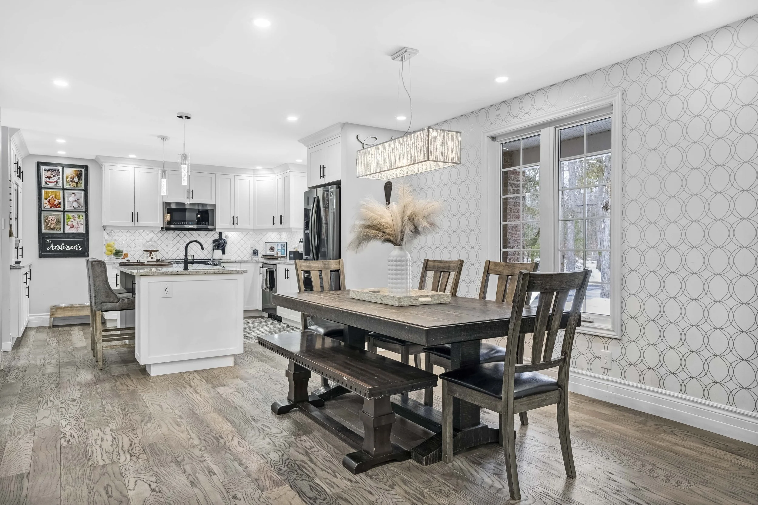 Ottawa Real Estate Photography  Open-concept kitchen and dining area with white cabinets, a wooden dining table with chairs, natural light from windows, and decorative wall paper.