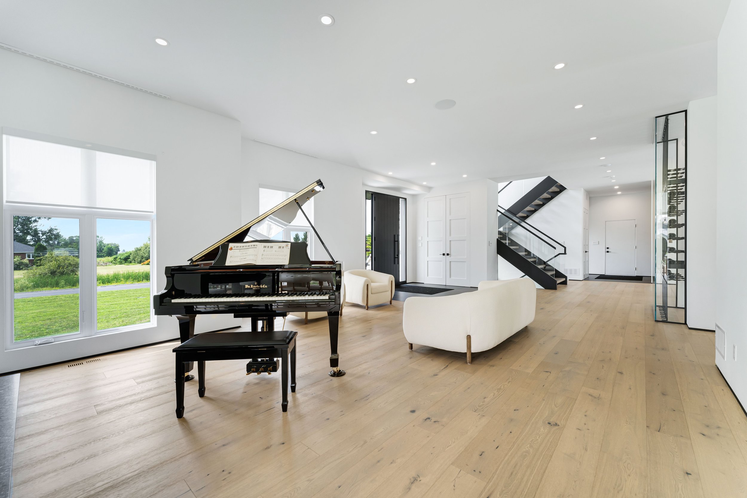 Modern living room with grand piano and white couches, featuring large windows, hardwood floors, and a staircase. Real estate photography ottawa