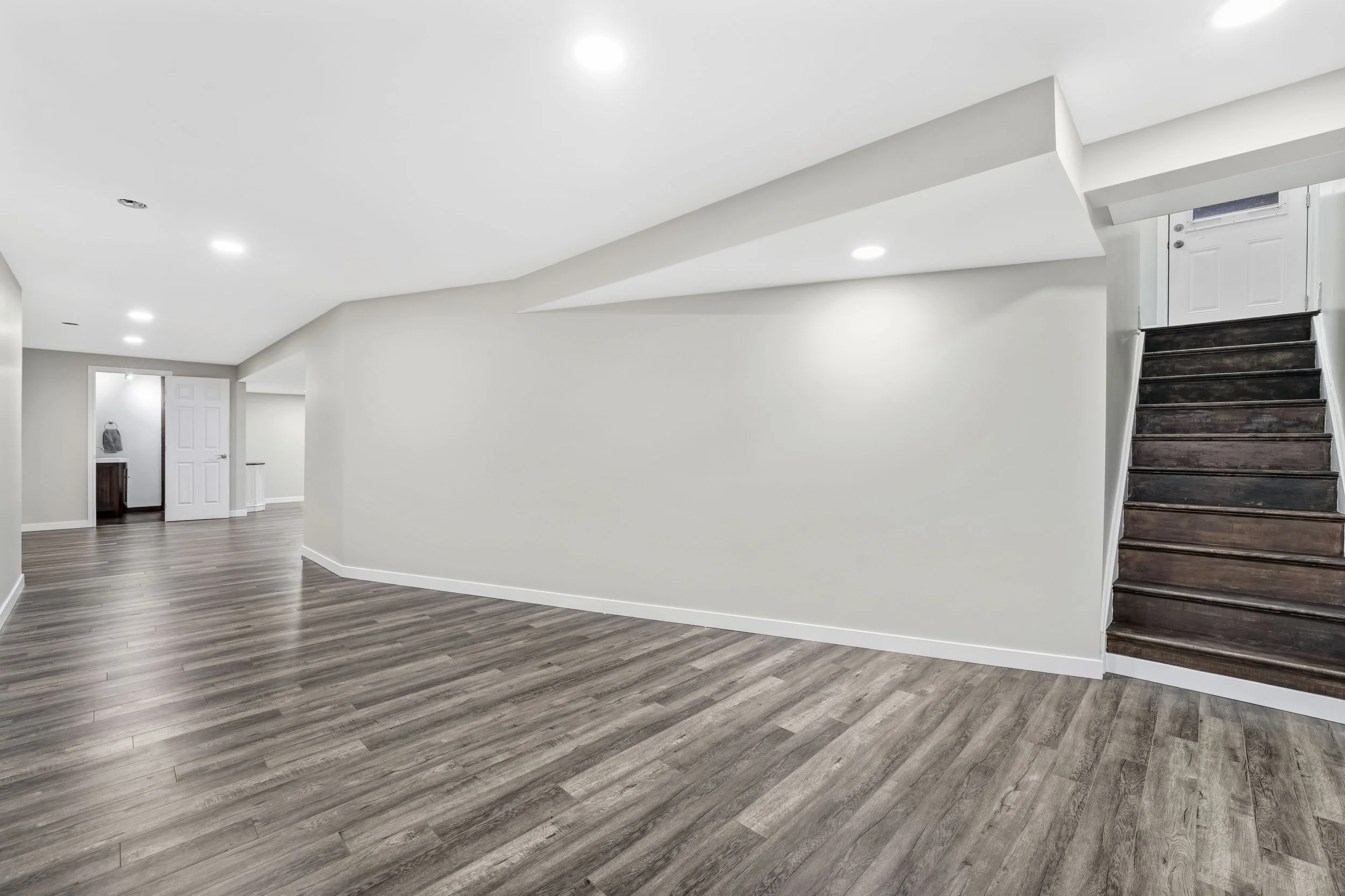 Ottawa Real Estate Photography  Empty basement room with white walls, dark wood flooring, and a staircase leading to a door at the top.