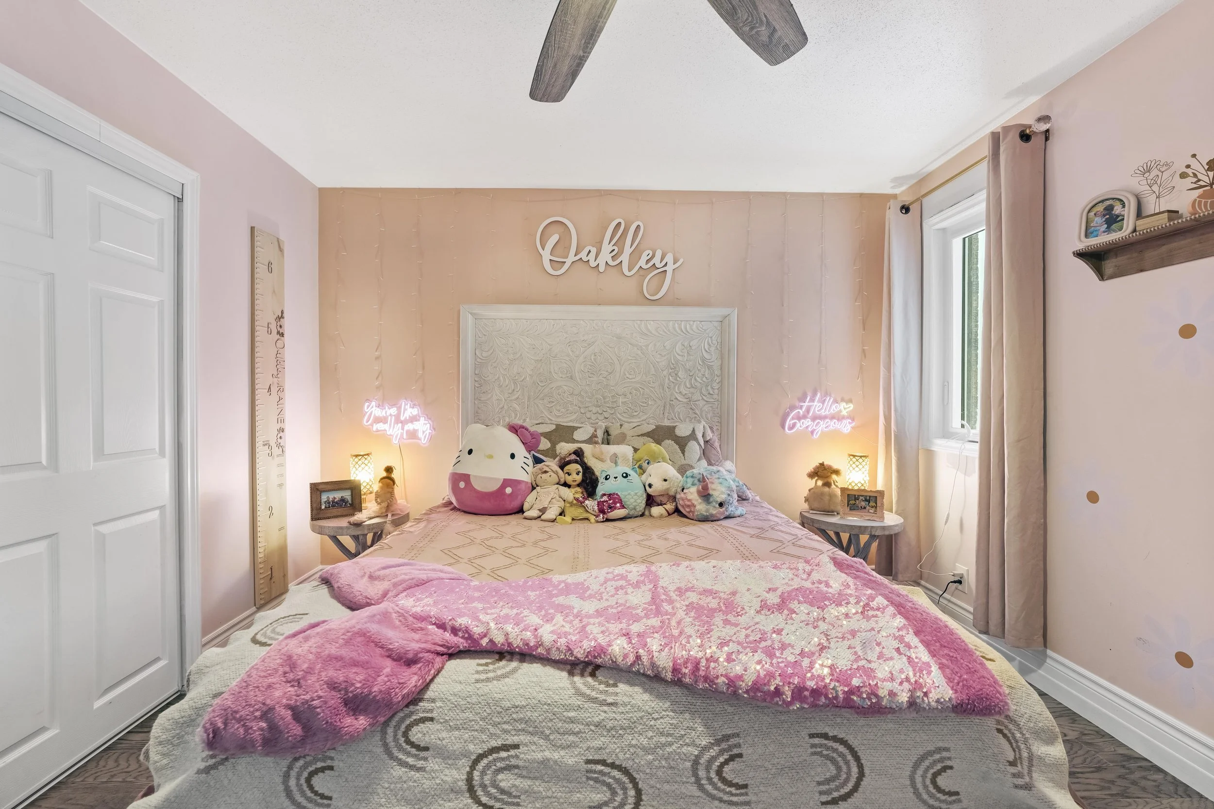A cozy bedroom with a bed covered in plush toys and dolls, soft pink blankets, and illuminated neon signs on the wall. The wall behind the bed is painted in a pastel peach shade with the name "Oakley" displayed above. There are two nightstands with l