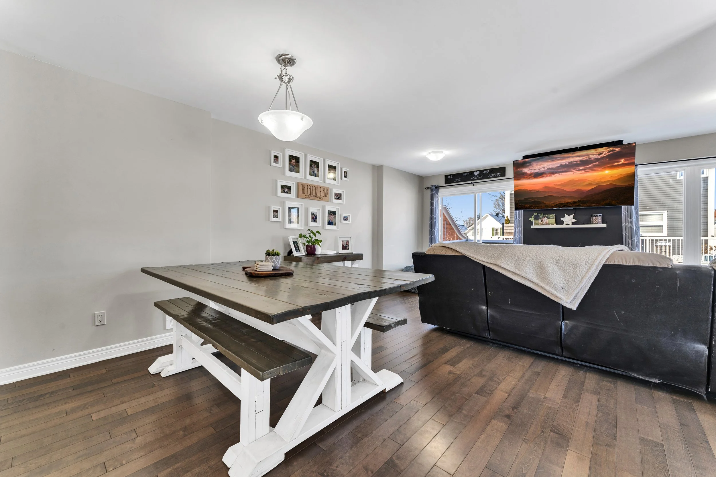 Living room with a dining area and TV, featuring a wooden dining table with bench seats, white walls, wall art, and a couch with a blanket, with large windows showing neighboring houses.
