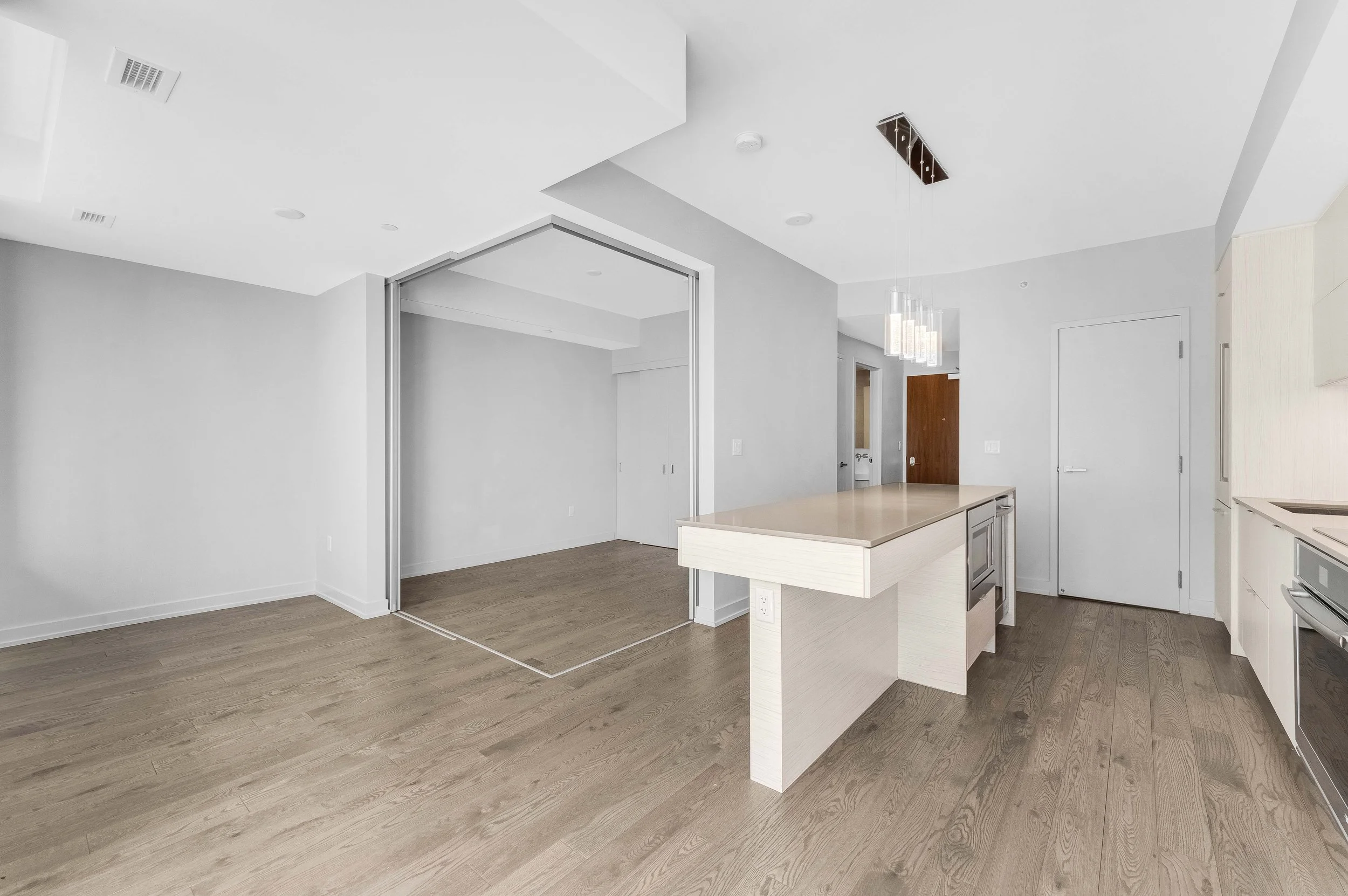 NV3D Ottawa Real Estate Photography Interior of a modern, minimalistic house with a kitchen and a living area. The space has white walls, wood flooring, and a ceiling with recessed lighting and a hanging light fixture.