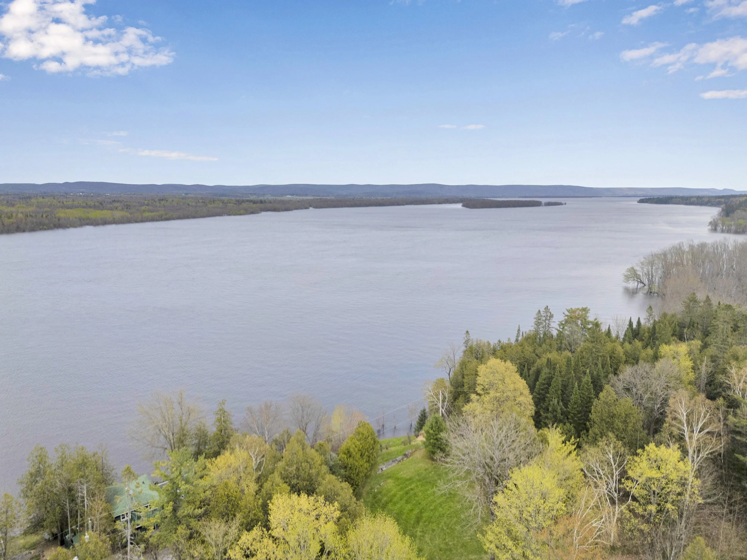 Ottawa Real Estate Media Services  An aerial view of a large river flowing through a landscape with lush green trees and hills in the background under a partly cloudy sky.