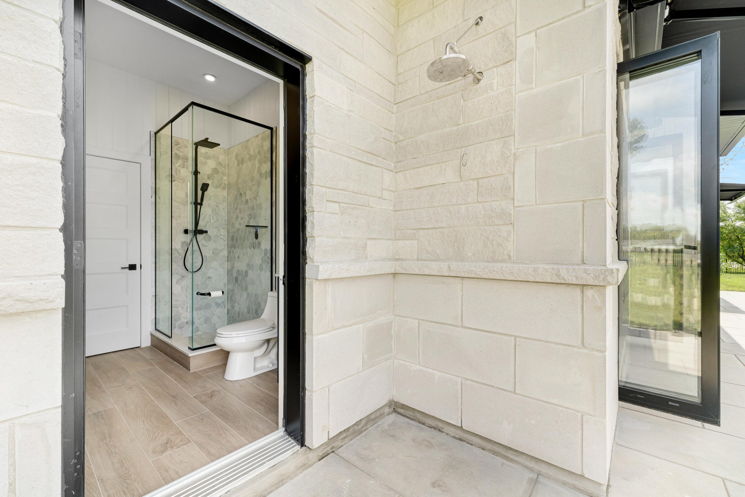 Outdoor shower area with nearby interior bathroom, featuring a glass-enclosed shower and toilet, set in light-colored stone walls and wood-effect tiles.
