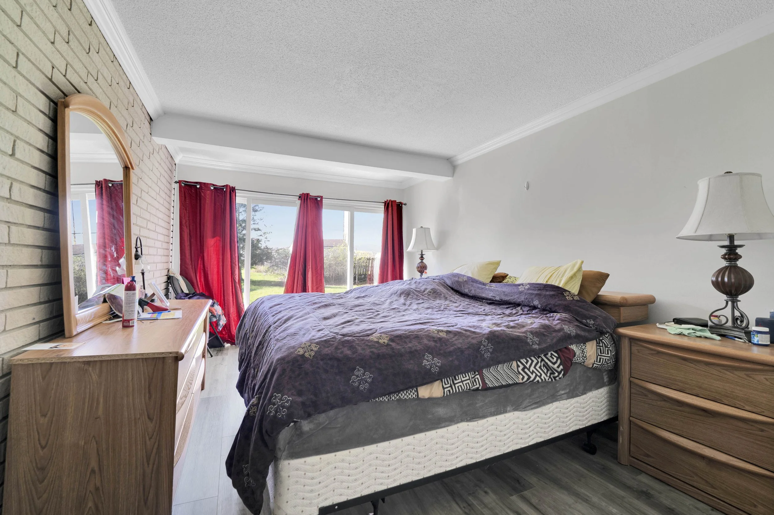 OA bedroom with a bed covered in a dark patterned quilt, with yellow pillows. There are red curtains on the windows, a wooden dresser and matching nightstand, and lamps on both sides of the bed. A mirror and various personal items are on the dresser.
