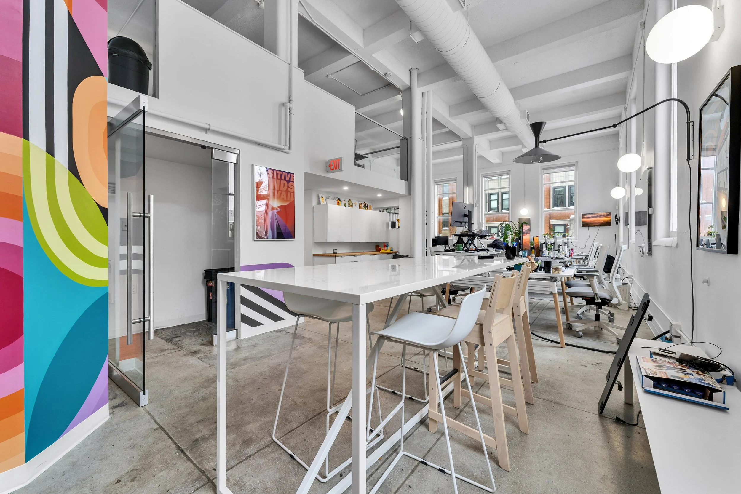Modern office with white walls and high ceiling, large windows, workstations with computers, white chairs, colorful abstract murals on the wall, and decorative plants.