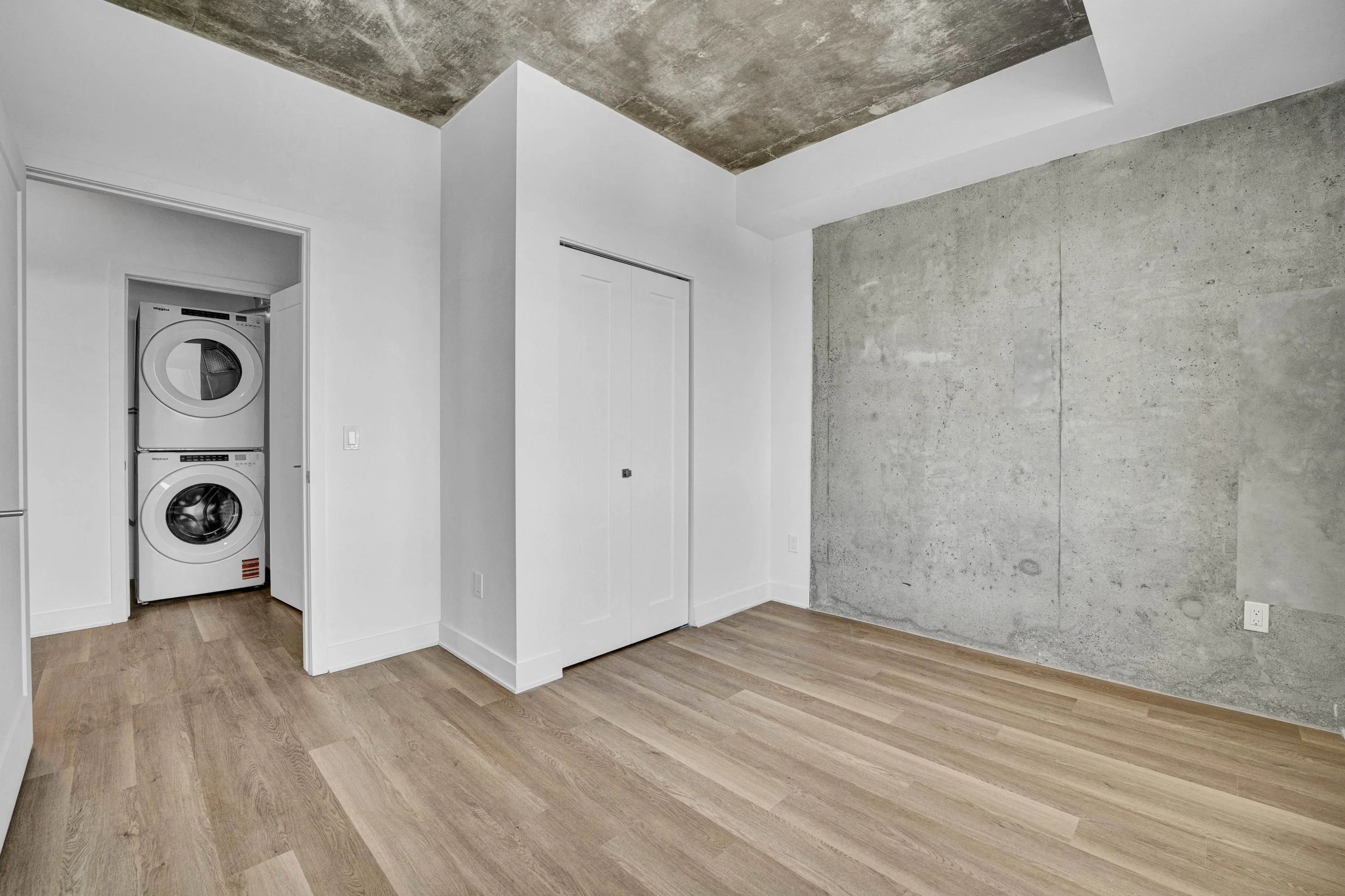 Interior room with wooden flooring, white walls, a concrete accent wall, and a small laundry area with a stacked washer and dryer.