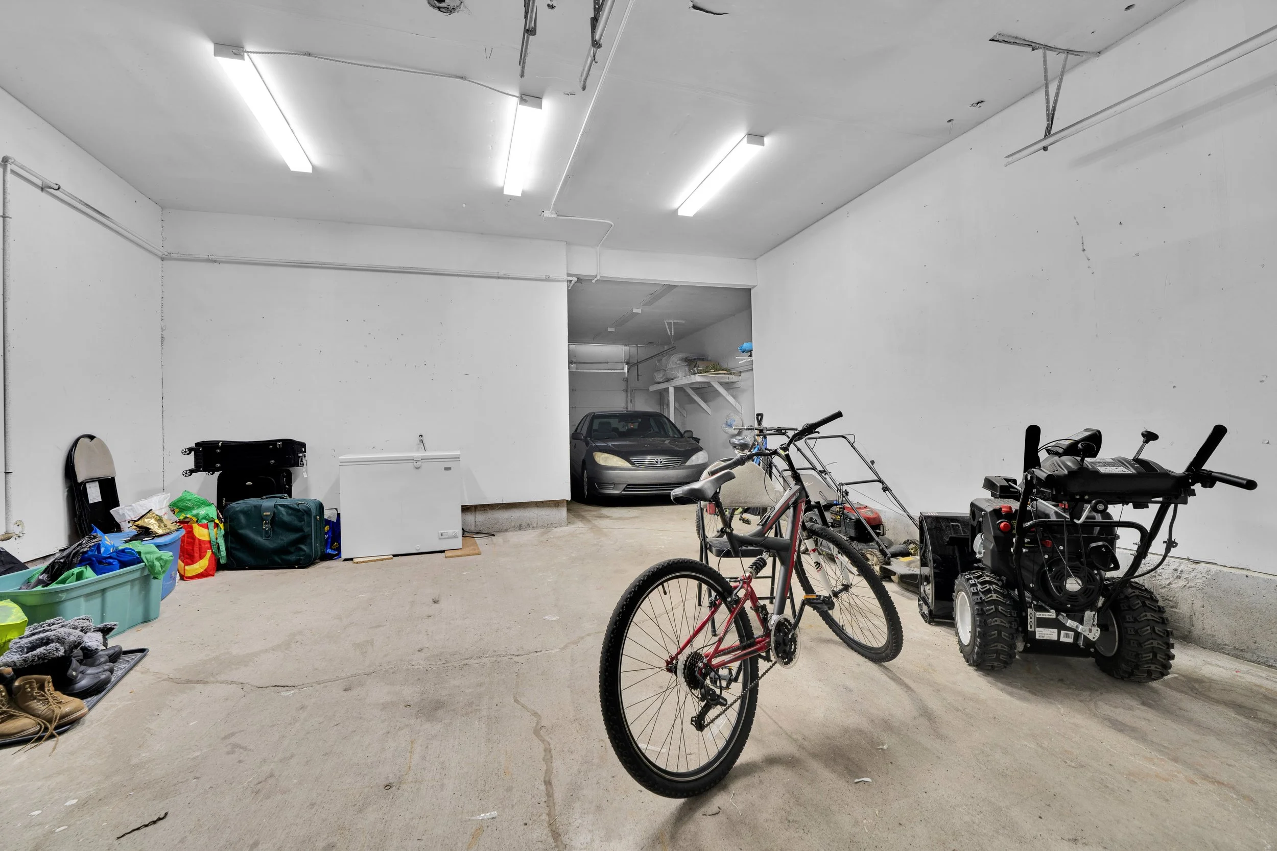 Ottawa Real Estate Media Services  A garage with bicycles, a toy tractor, a black car, and storage bins. The garage has white walls and a concrete floor, with fluorescent lighting and some items stored on shelves and the floor.