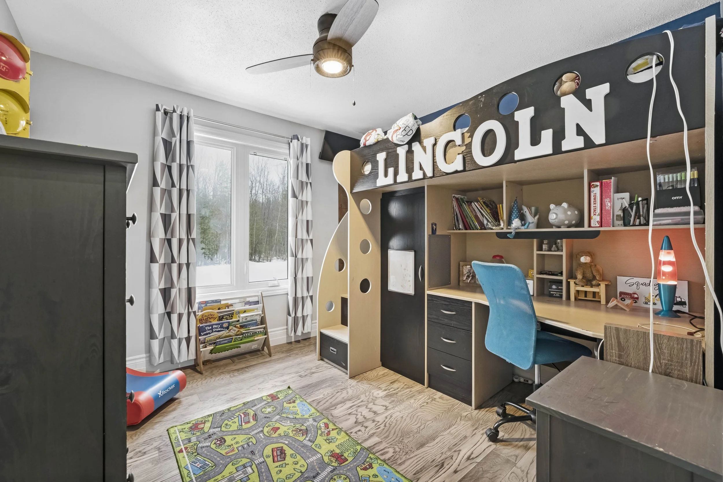Child's bedroom with a window, patterned curtains, a colorful car-themed rug, a black dresser, and a custom wooden loft bed with the name 'Lincoln' on top and a reading nook underneath. The room has a ceiling fan and a small bookshelf near the window