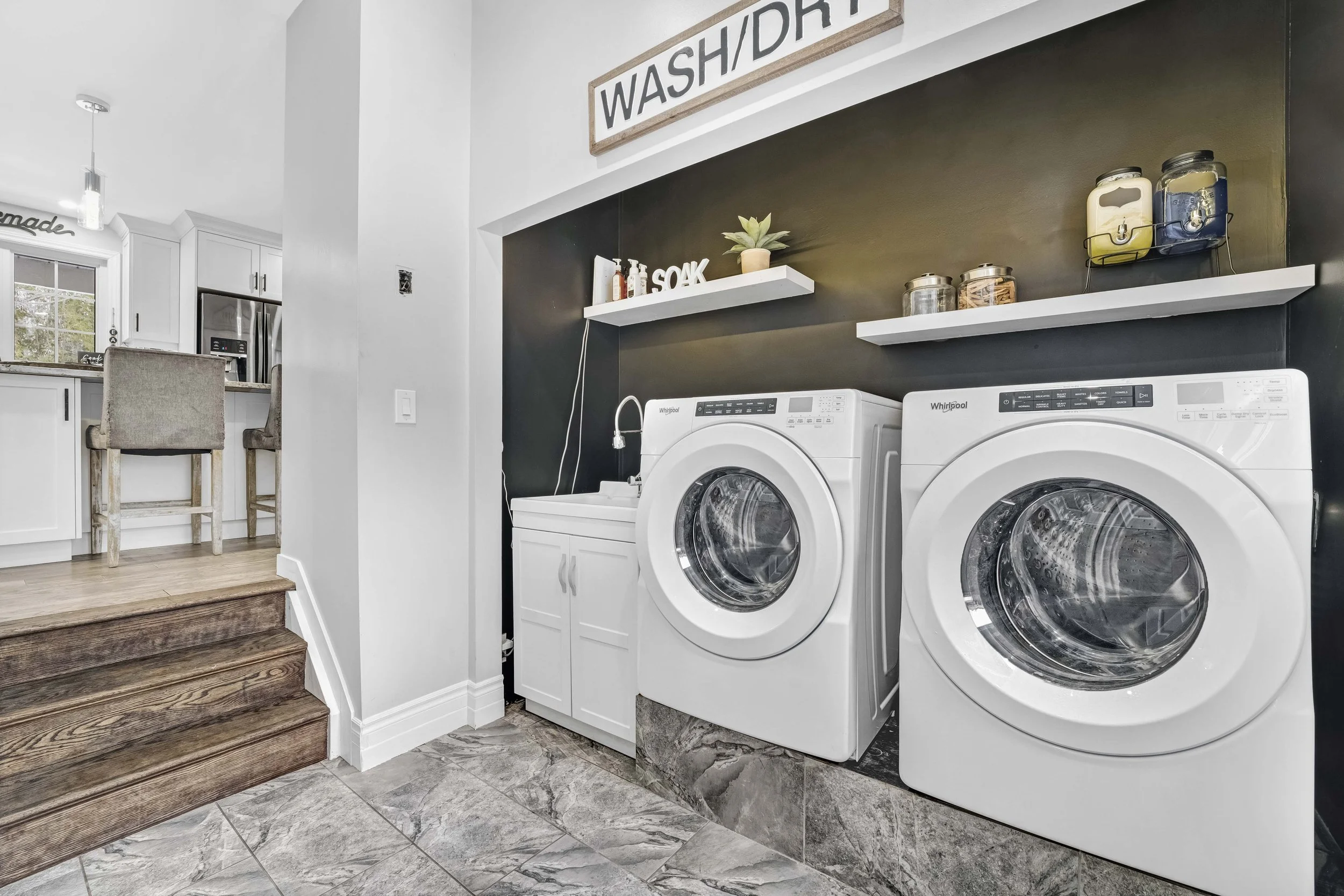 photographie immobiliere outaouis  Laundry room with a washer and dryer, black wall with white shelves, small cabinet with sink, and decorative items, partially open to kitchen area with barstools and window.