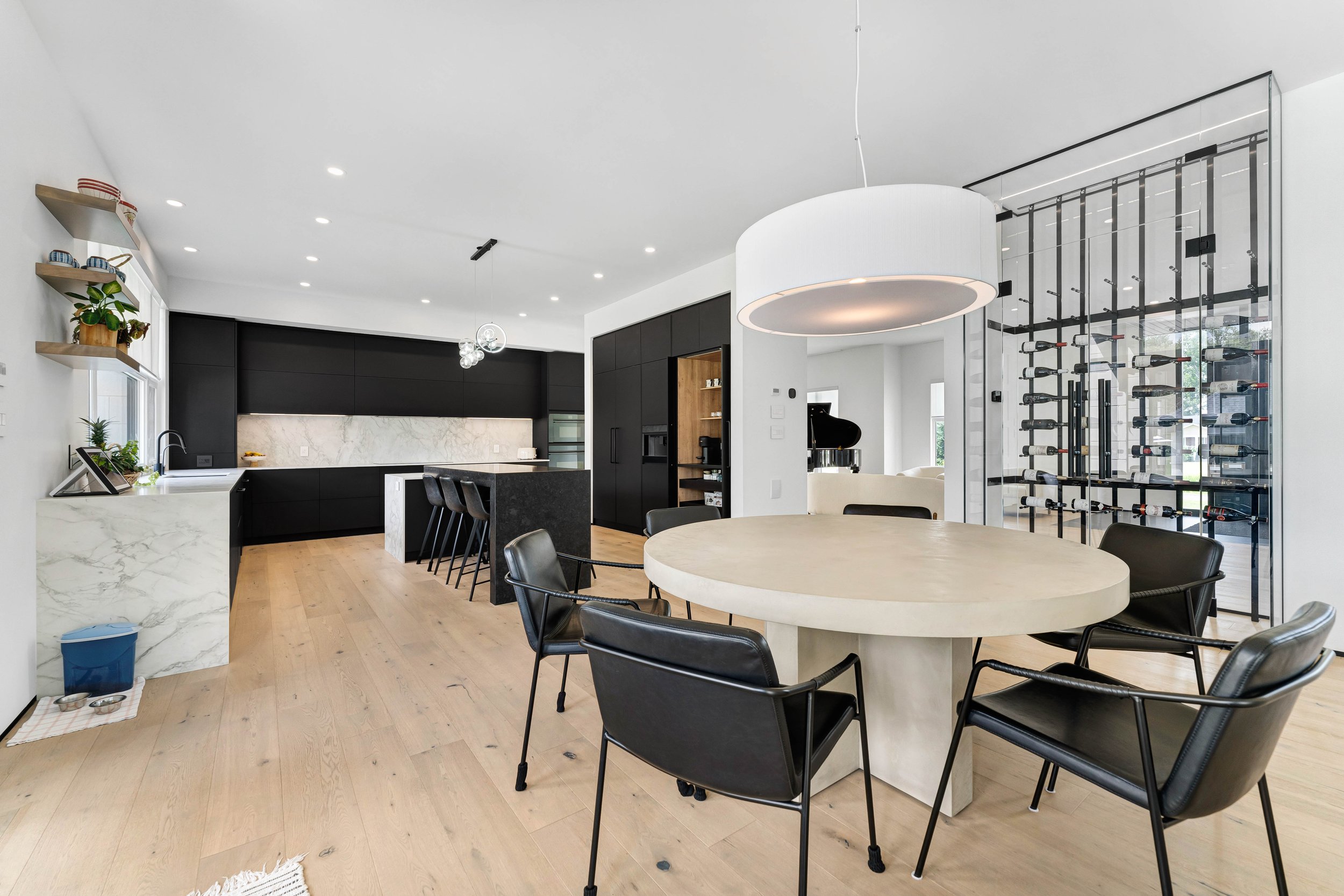 Modern kitchen and dining area with black cabinetry, marble countertops, light hardwood flooring, round dining table with black chairs, large white pendant light, and wine rack in the background.