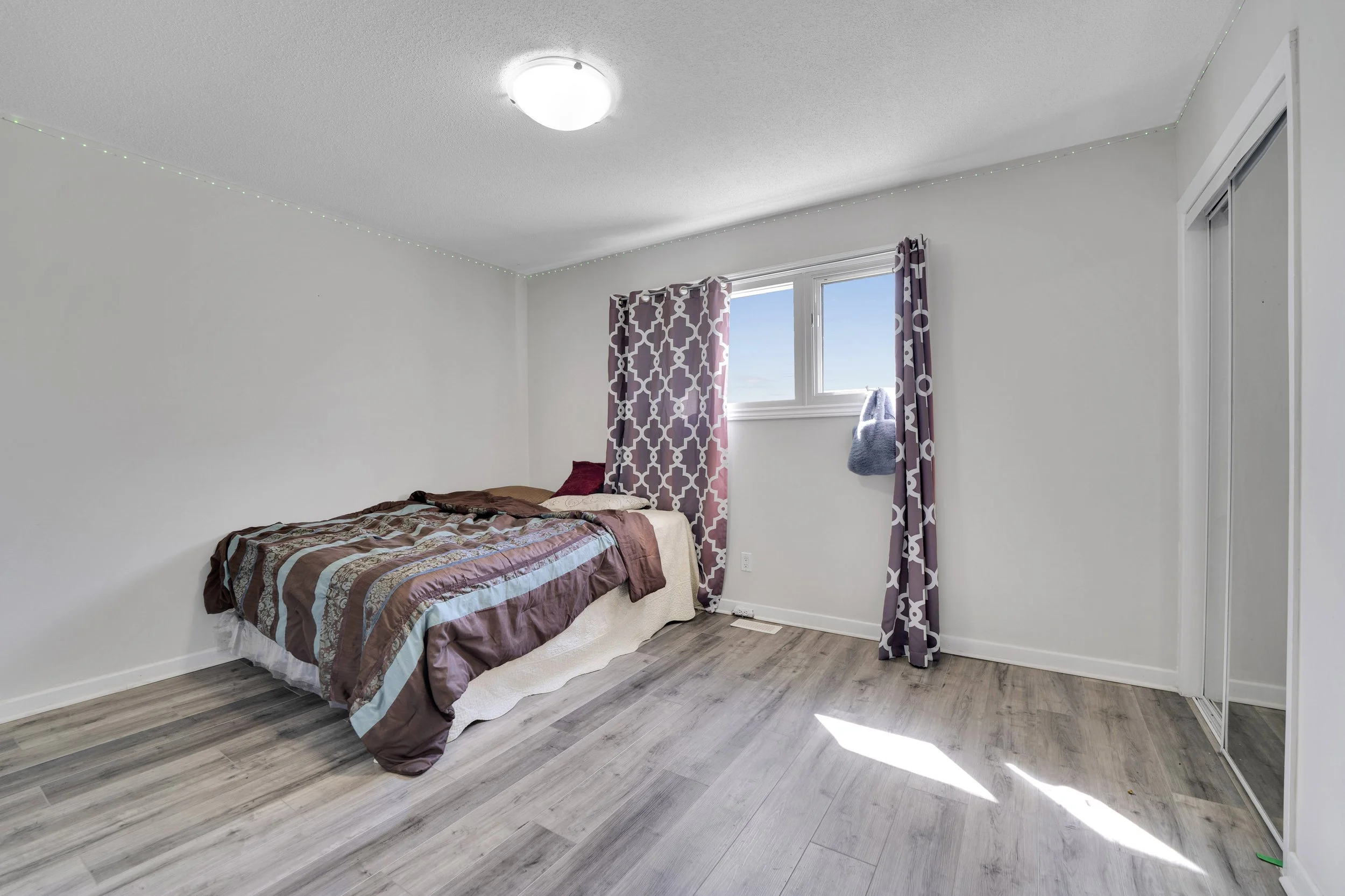 A bedroom with a bed covered in a patterned quilt, purple and brown pillows, lavender curtains with geometric patterns, and a window showing a clear blue sky. The room has white walls, a ceiling light fixture, wood flooring, and a closet with sliding