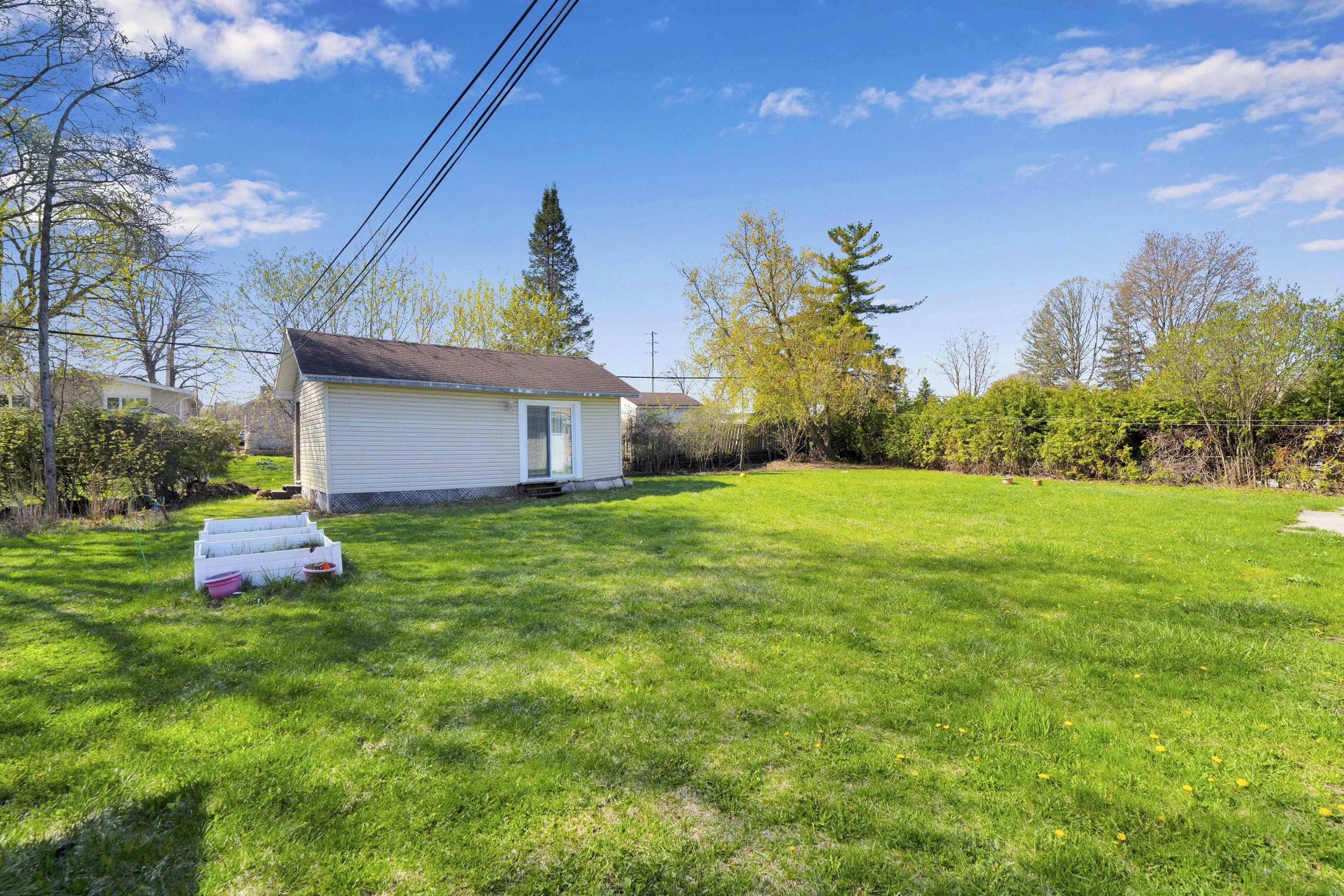 Ottawa Real Estate Media Services  A backyard with green grass, a small white house with a sliding glass door, trees with budding leaves, and a bright blue sky with some clouds.