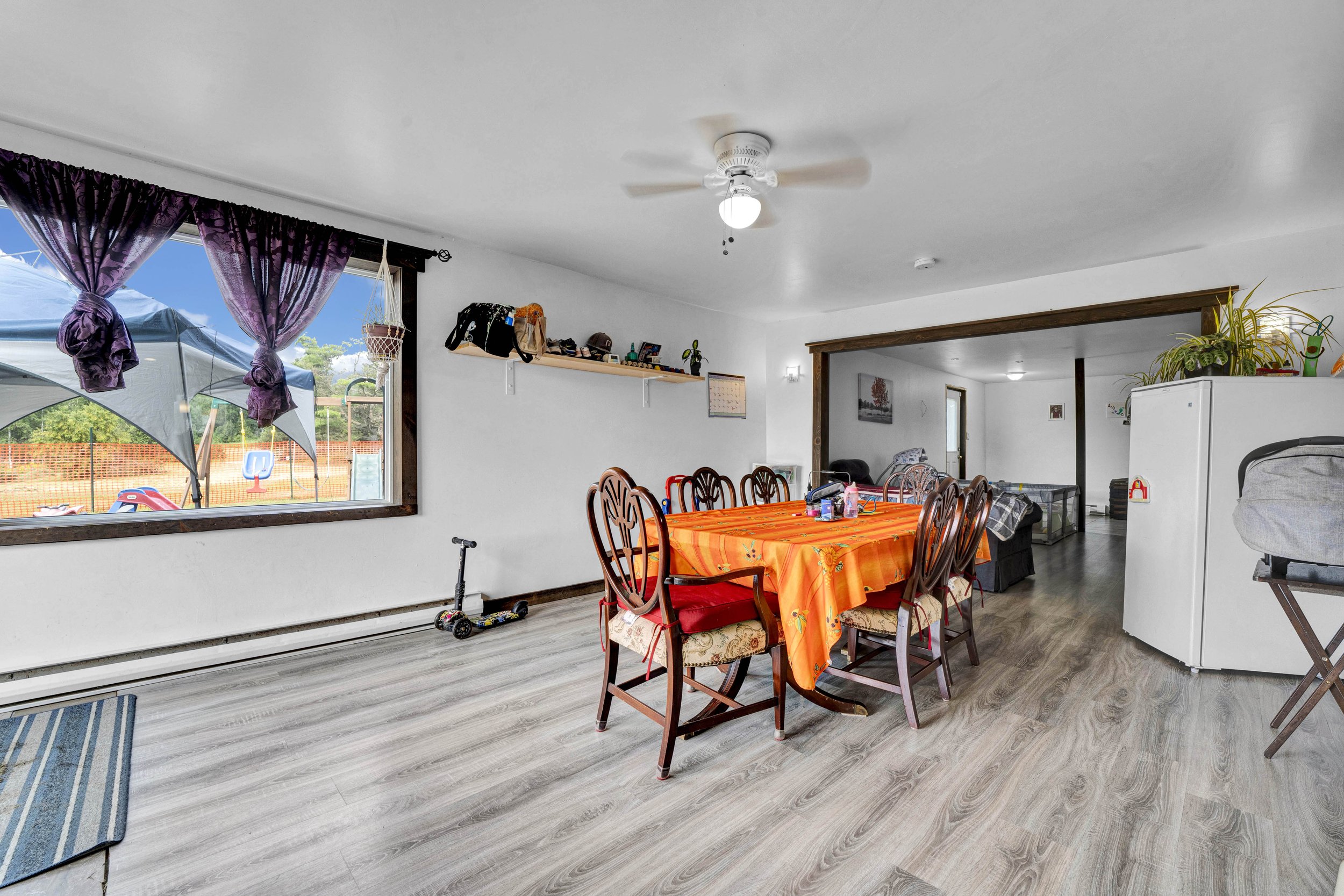 Interior of a dining area with a wooden table covered by an orange tablecloth, surrounded by wooden chairs with red cushions. Large window with purple curtains shows an outdoor patio with umbrellas. Items such as backpacks, children's toys, and house