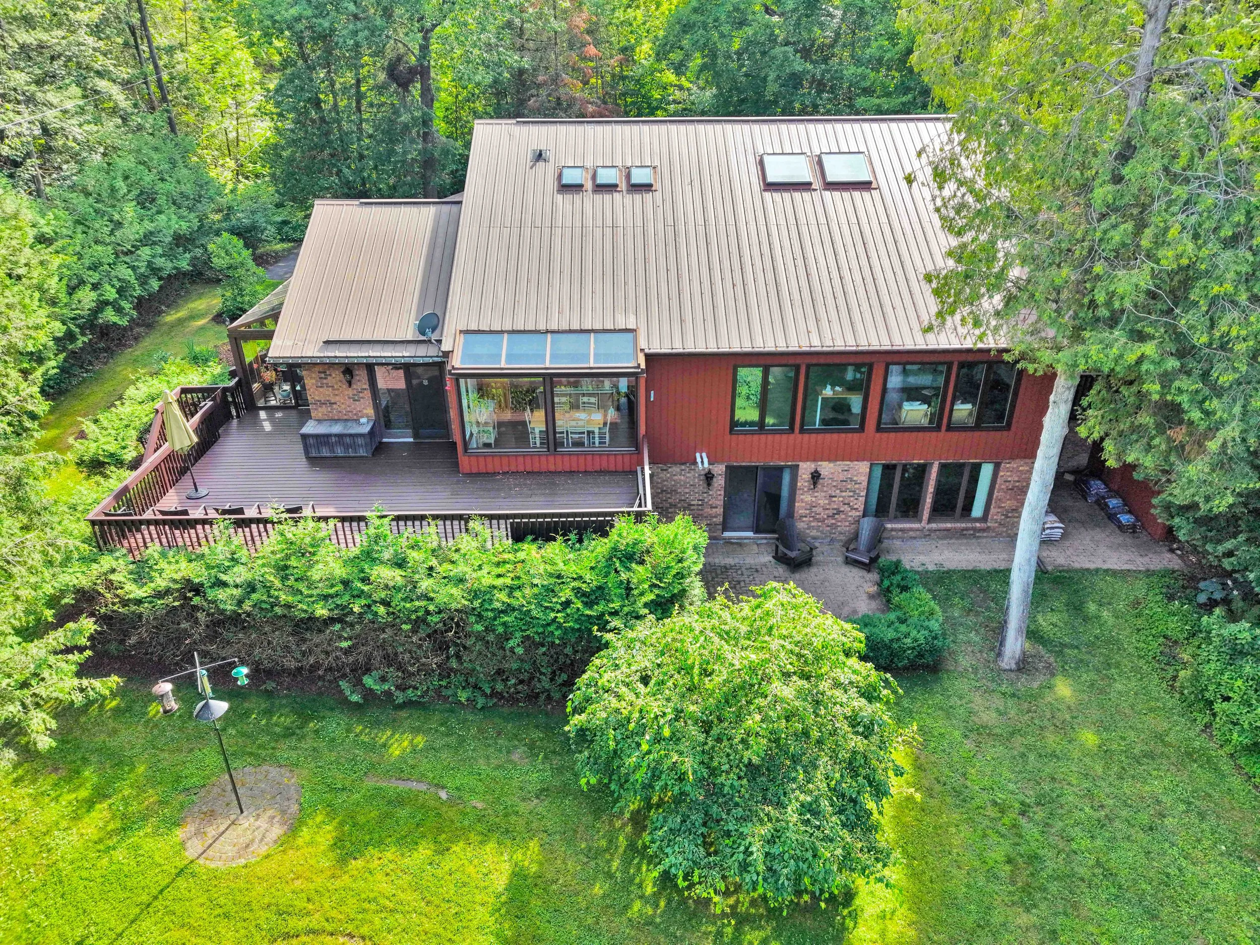 NVISION 3D Real Estate Media  An aerial view of a house surrounded by trees and greenery, featuring a large deck with outdoor furniture and a roof with skylights and green landscaping.