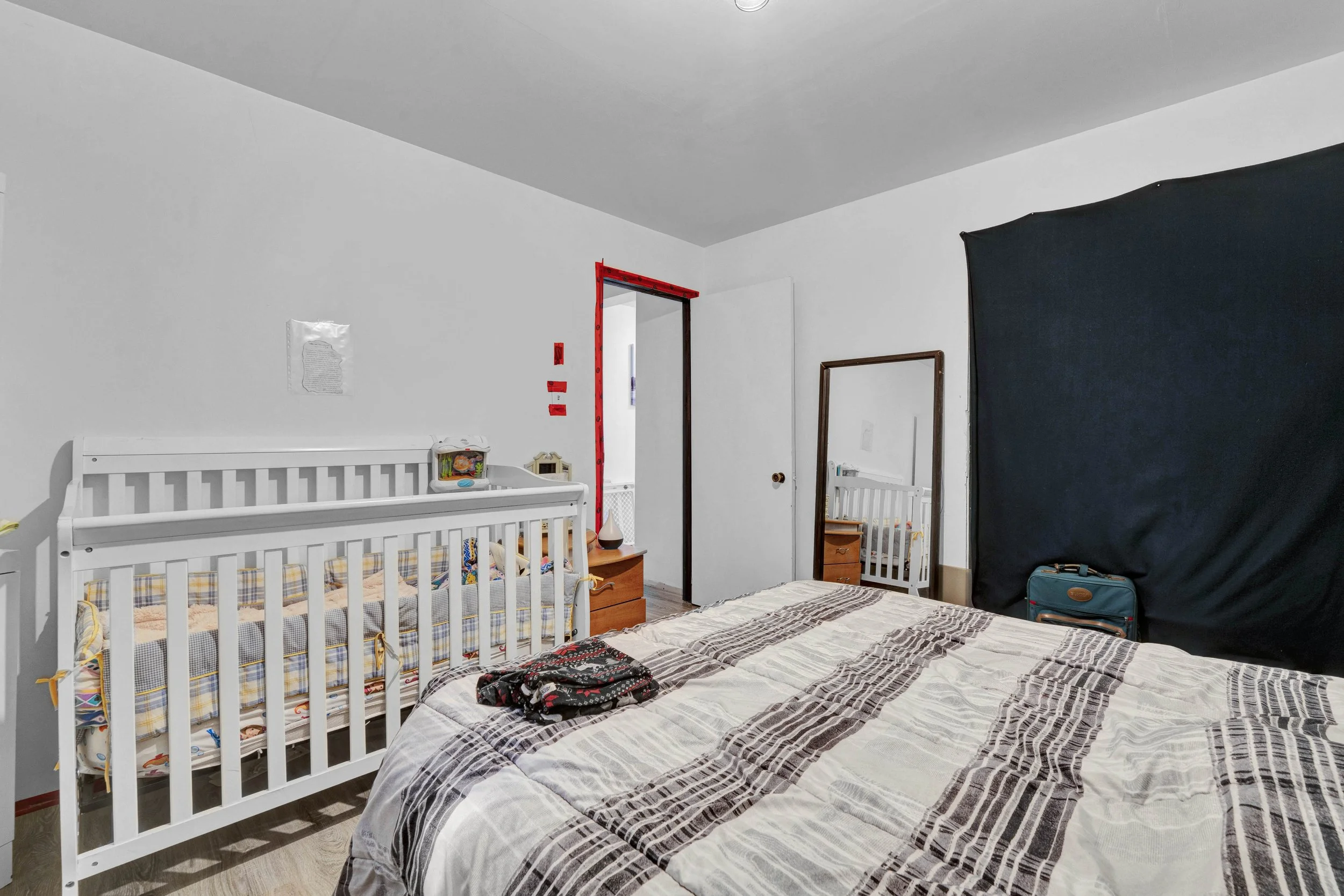 A bedroom with a large bed covered in a multi-patterned bedspread, a white baby crib with a colorful mattress and bedding, a small wooden nightstand, a standing mirror, and a dark privacy screen. The walls are plain white, with a door and some red ta