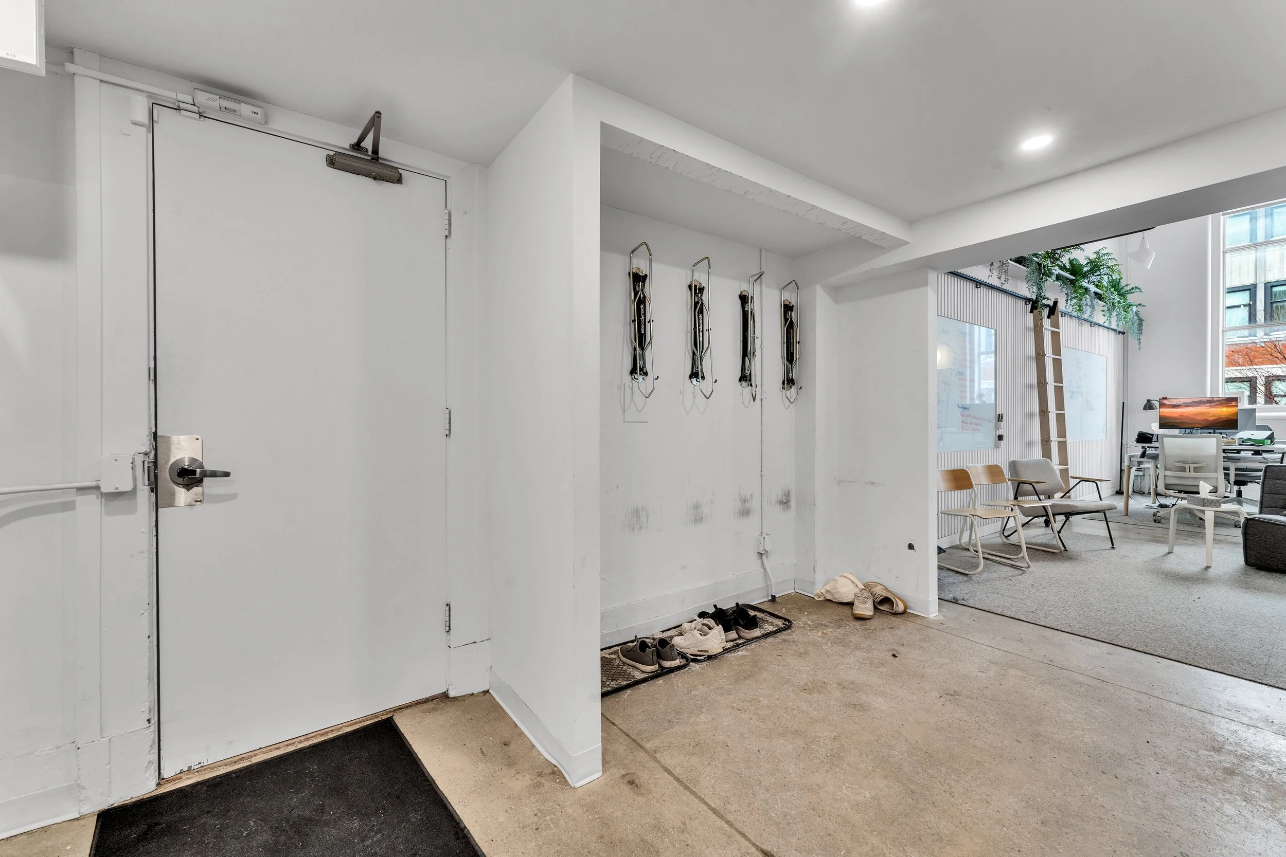 Interior of an office with white walls, a white door with a handle lock, wall hooks with hanging cords or straps, and several pairs of shoes on the floor. In the background, there is a workspace with chairs, a ladder, and a large window with plants a