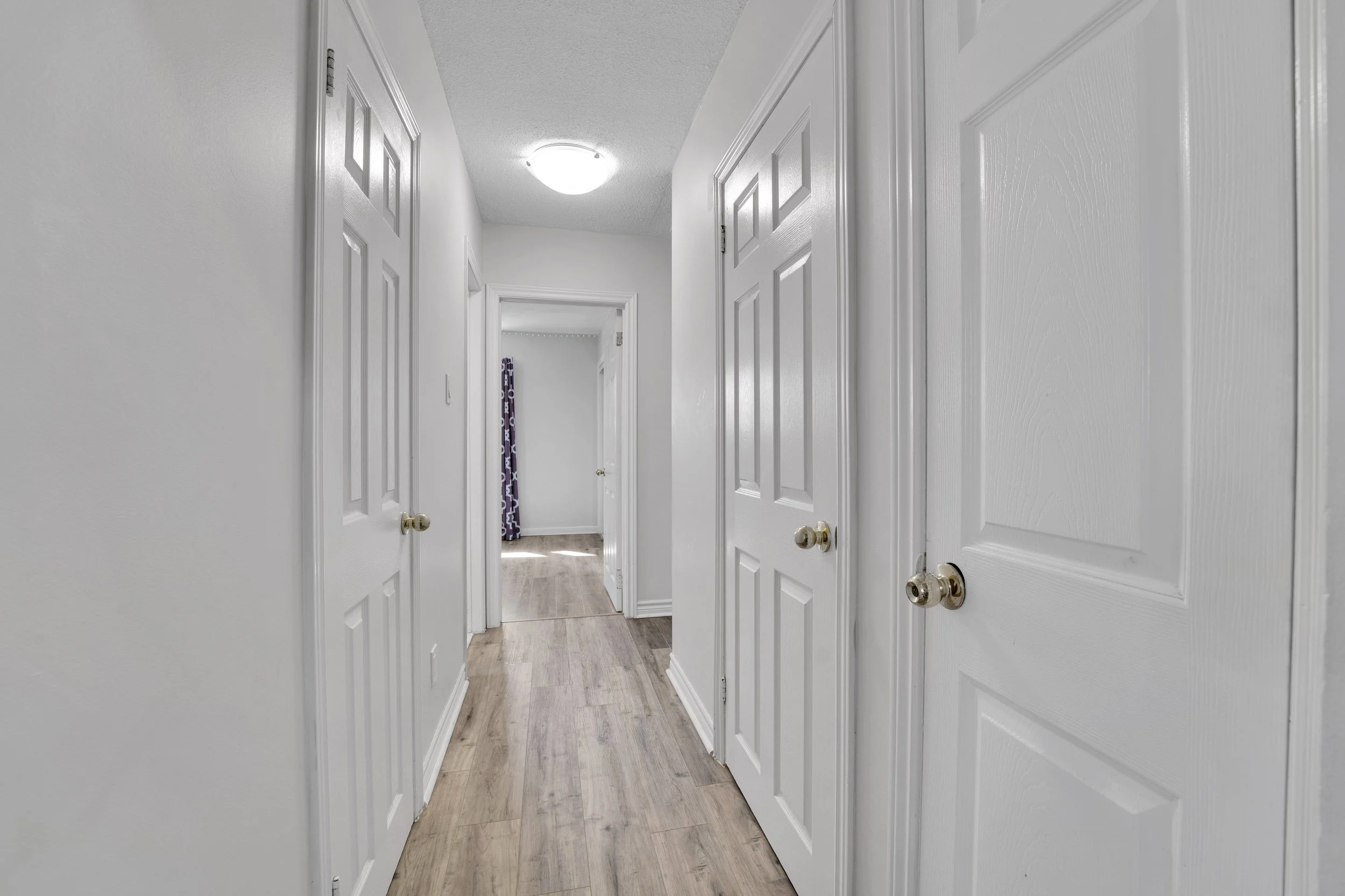 Ottawa Real Estate Media Services  A hallway with white walls and multiple closed white doors, leading to a room with a window covered by a purple curtain, and a ceiling light at the end of the hallway.