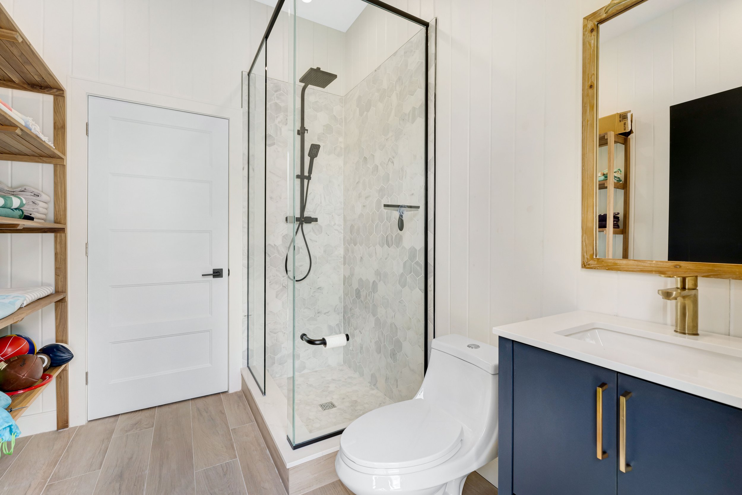 Modern bathroom with a glass shower enclosure, white tiles, wood shelving, a white toilet, and a navy blue vanity with a gold faucet and mirror.