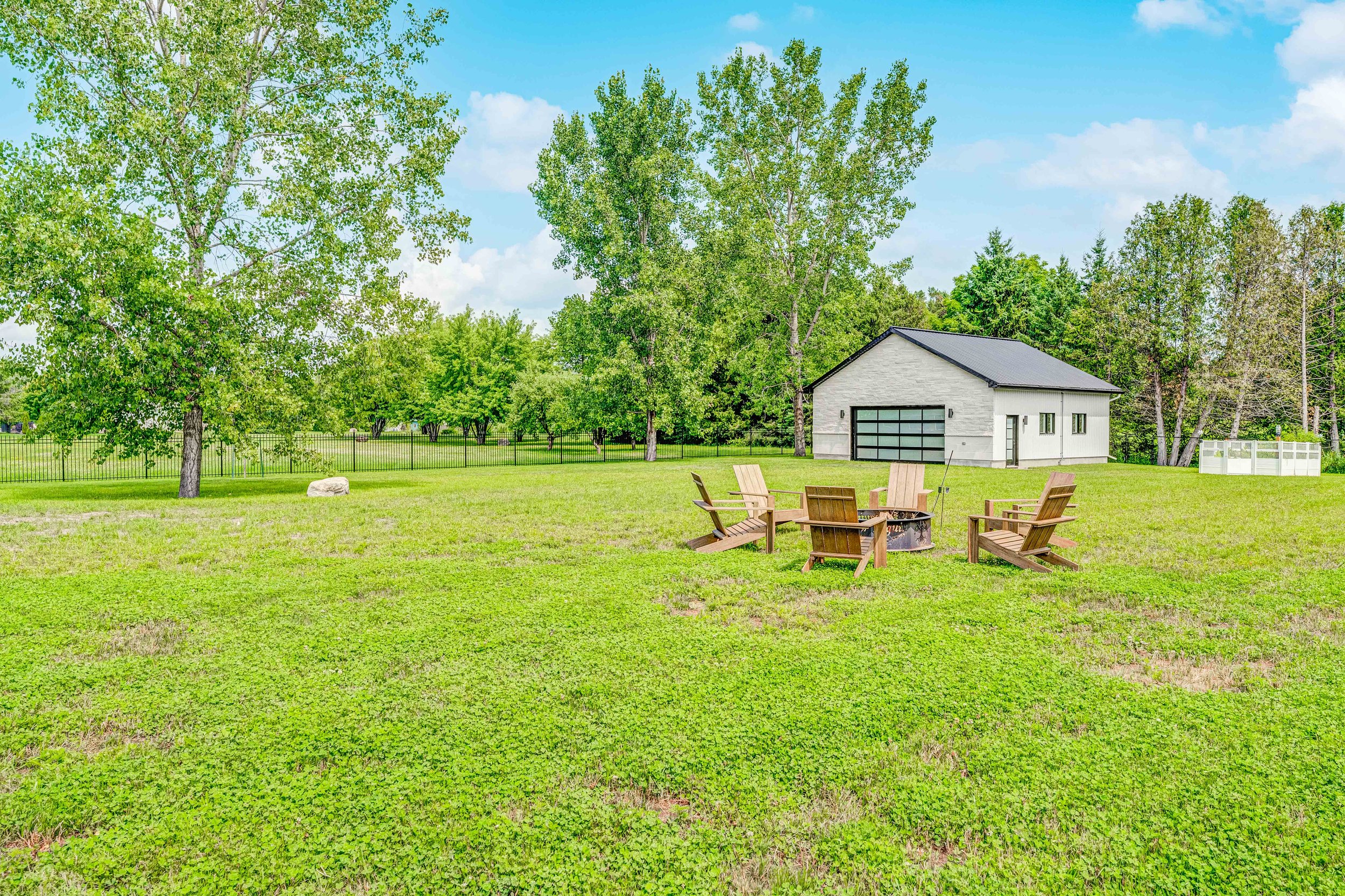 Open grassy backyard with a fire pit and wooden Adirondack chairs, surrounded by trees, with a modern detached garage in the background.