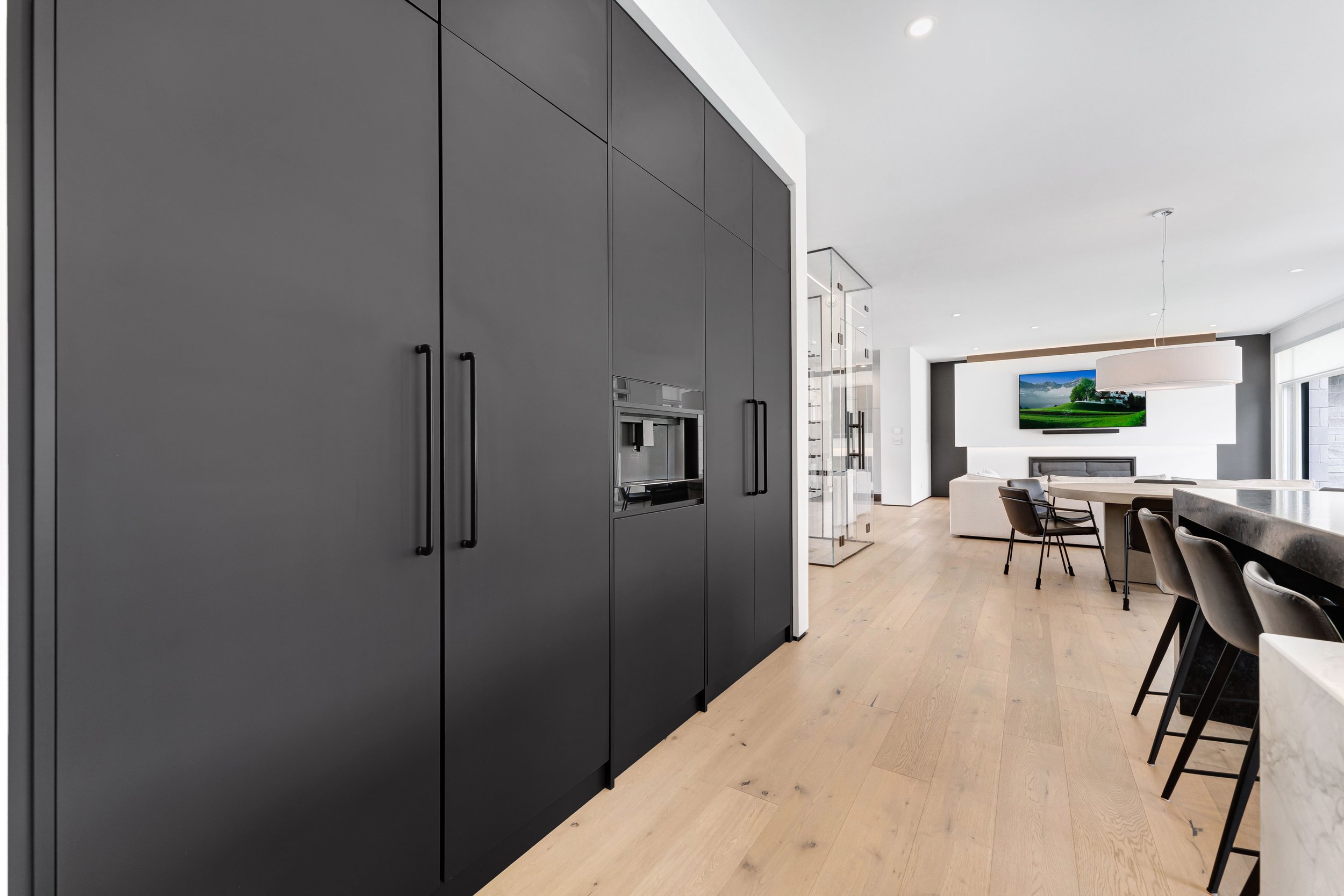 Modern kitchen interior with black cabinets, light wood flooring, bar stools, and a dining area with a mounted TV.
