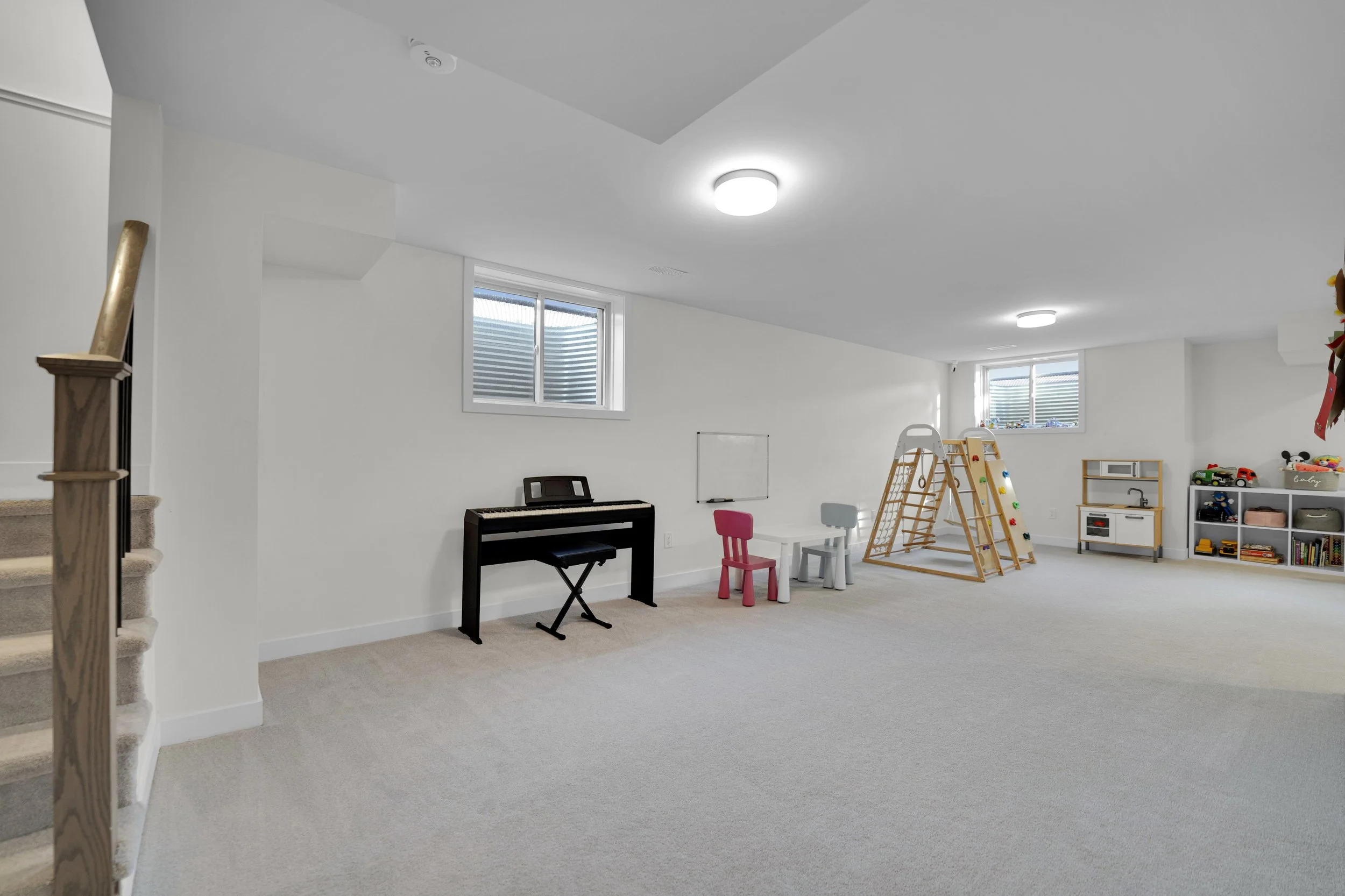 NVISION3D Real Estate Photography Ottawa  A spacious basement playroom with white walls and ceiling, featuring a piano, small chairs, a ladder, and toy storage units. Two windows provide natural light, and ceiling lights illuminate the room.