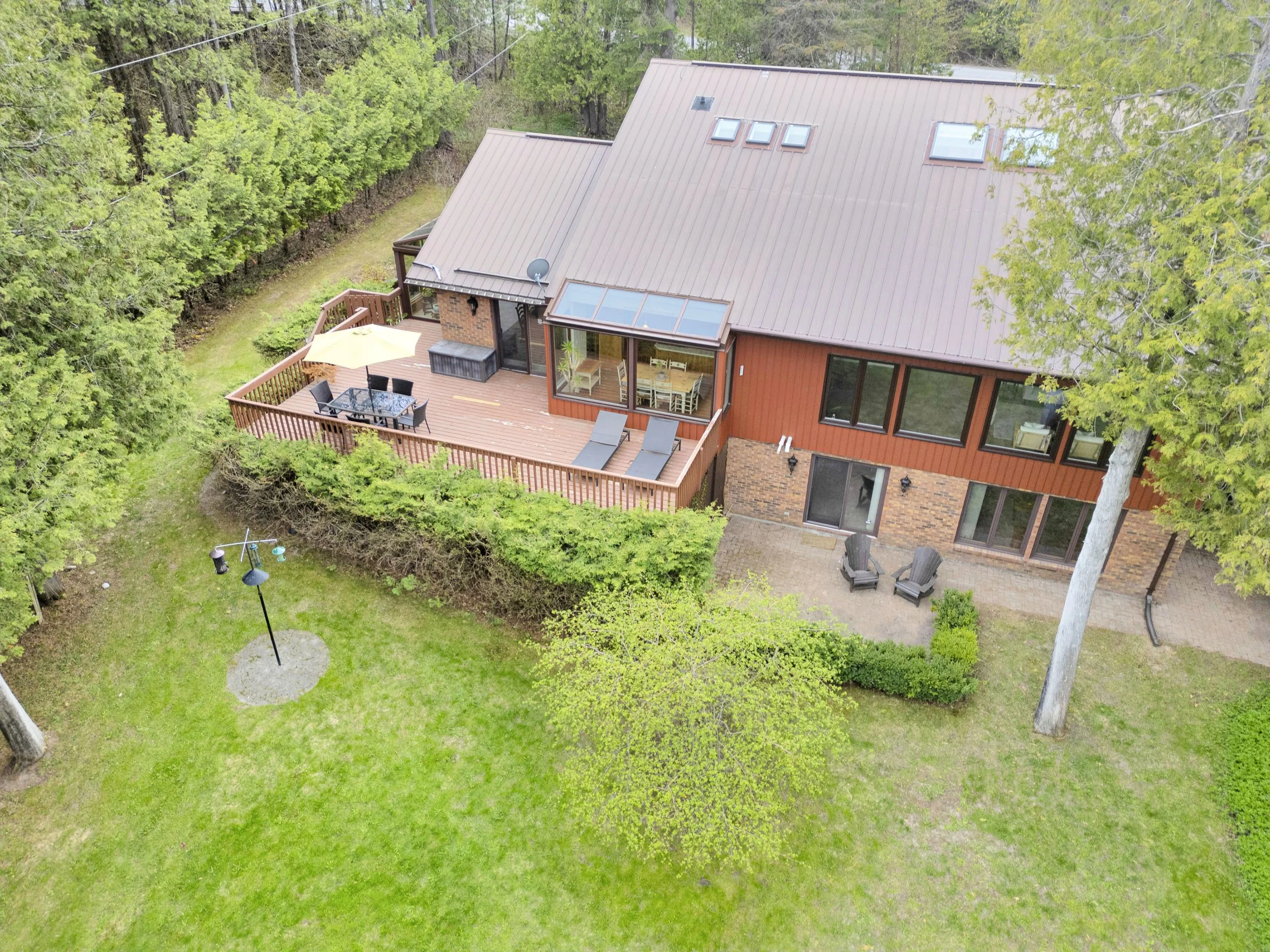 Ottawa Real EstAerial view of a two-story house with a brown metal roof, a wooden deck with patio furniture including a table, chairs, and a yellow umbrella, surrounded by green trees and a lush yard with a small garden path and decorative lamp post.