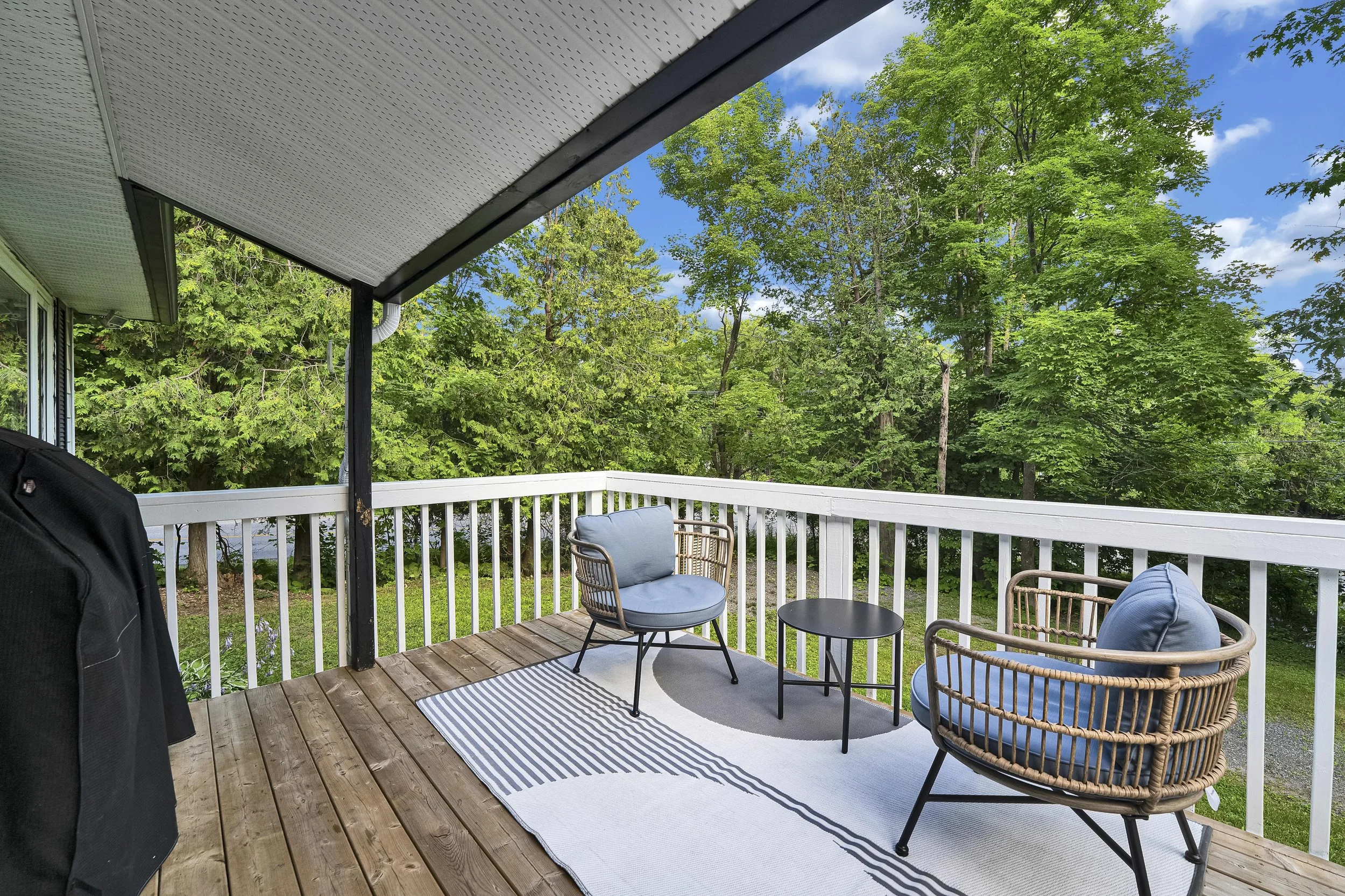 Ottawa Real Estate Photographer Back porch with outdoor furniture, including two round chairs with blue cushions, a black table, and a striped outdoor rug, overlooking a green wooded yard and blue sky with clouds.