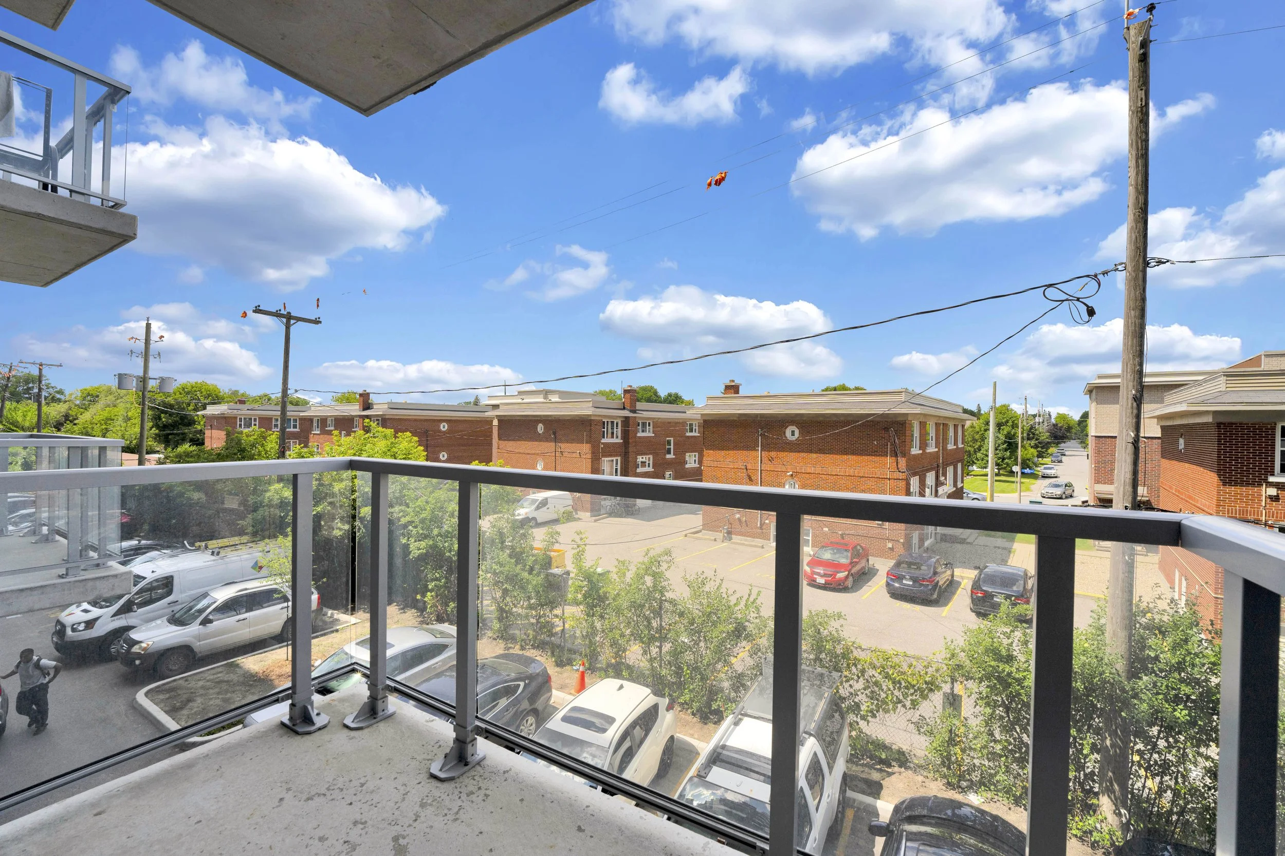 Ottawa Real Estate NVISION 3D  View from a balcony showing parking lot, trees, brick apartment buildings, power poles with wires, and a bright blue sky with scattered clouds.