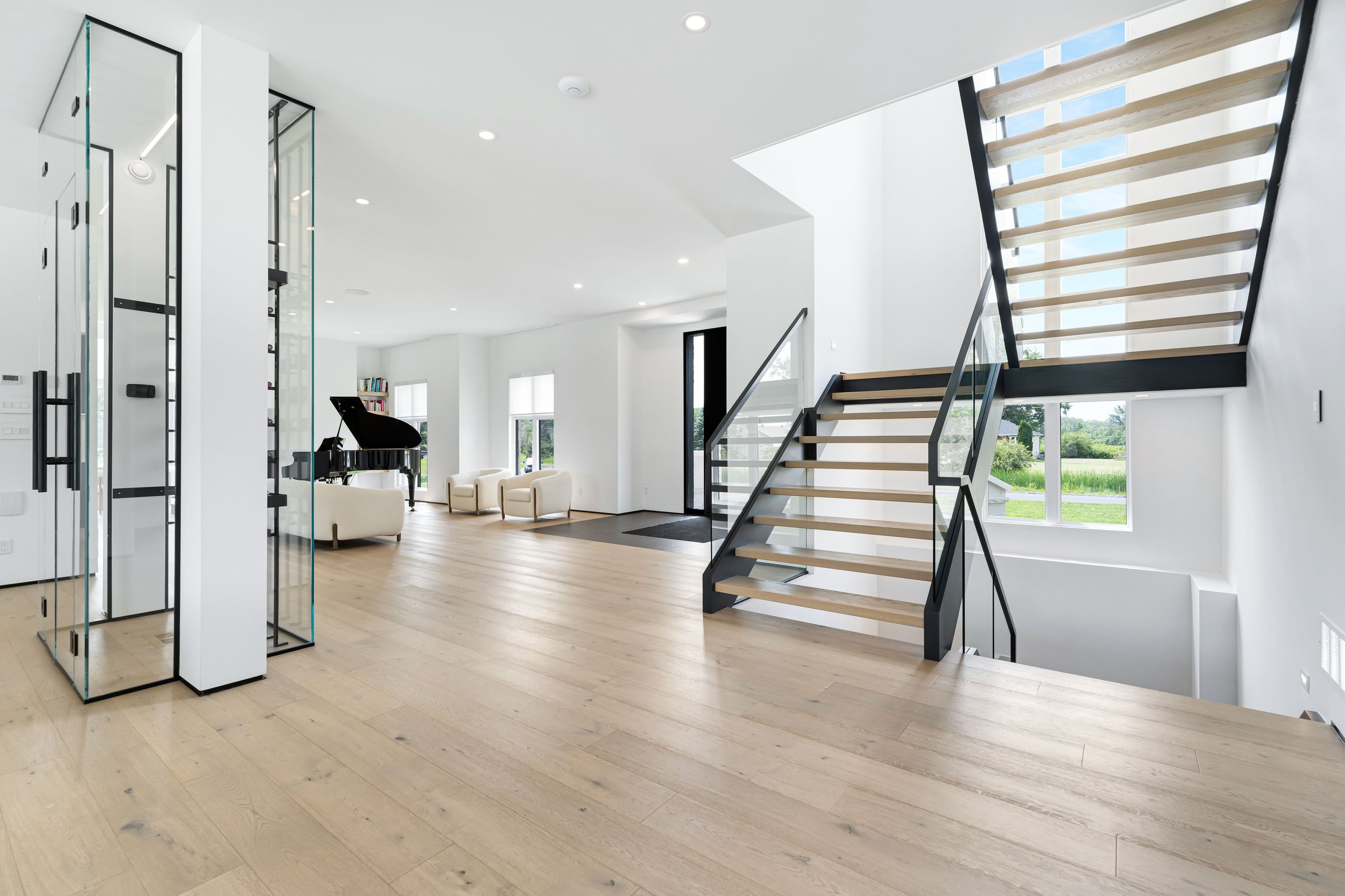 Modern interior with wooden flooring, open staircase, glass elevator, grand piano, and white seating area.