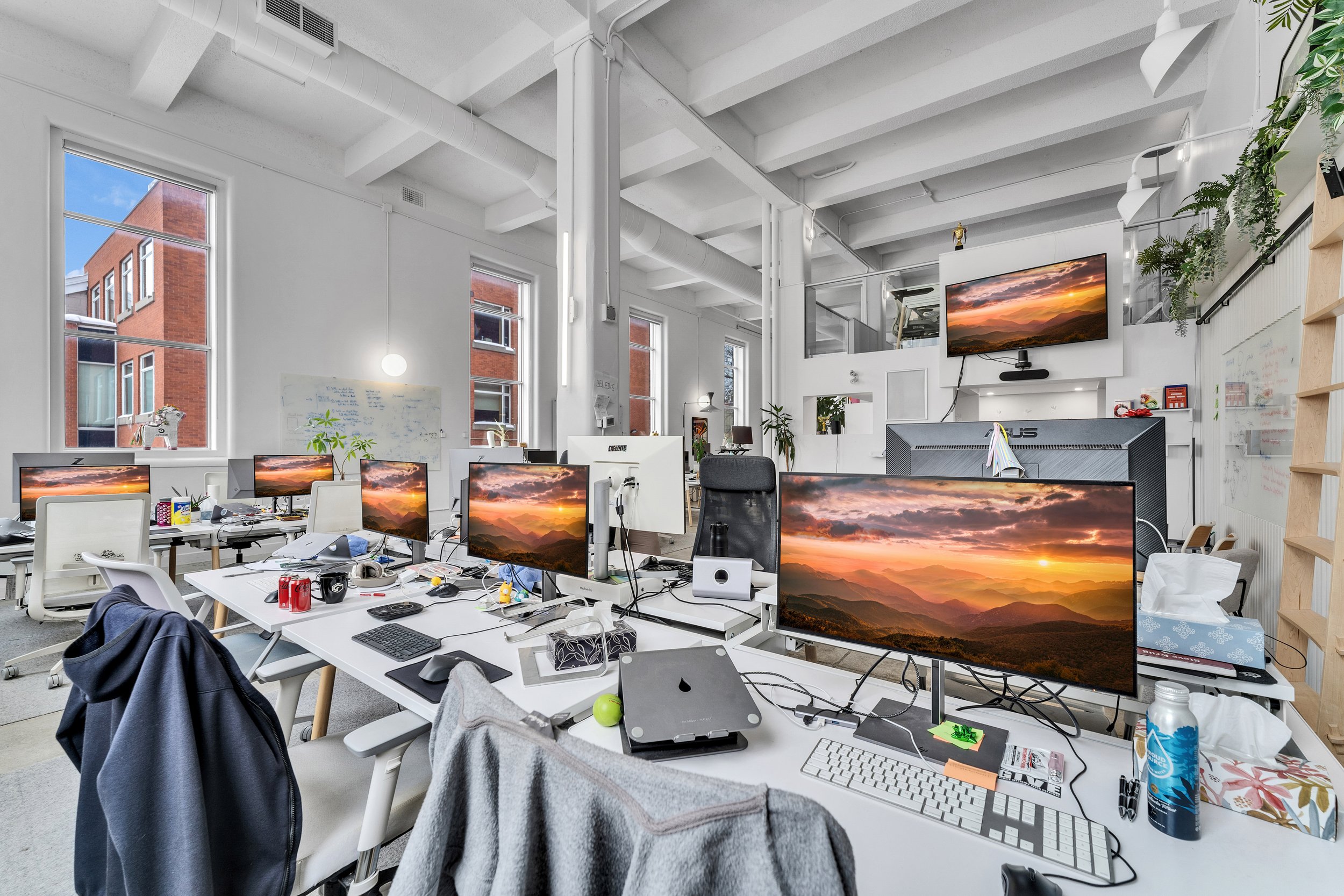 Open office workspace with multiple desks, computer monitors displaying a sunset mountain scene, office chairs, paper tissues, and personal items. Windows show neighboring red brick buildings.