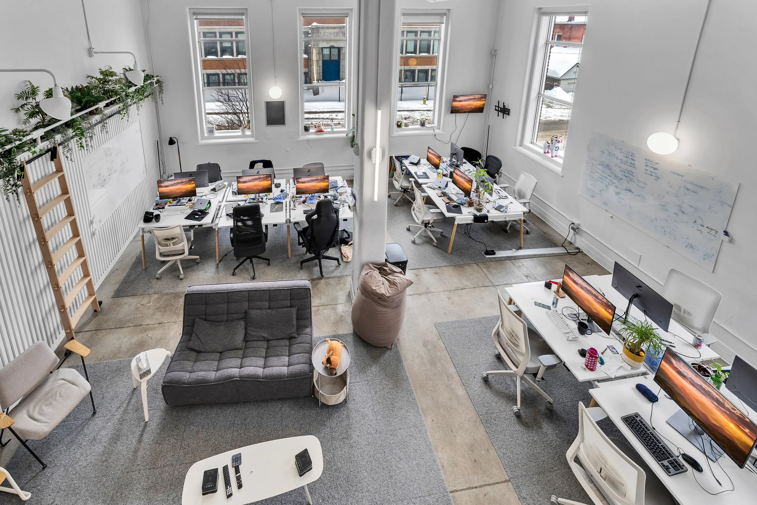 Open-plan office with multiple desks and chairs, computer monitors displaying sunset images, books, plants, whiteboard with writings, gray sofa, and a white table with remotes, in a room with large windows showing a snowy outdoor scene.