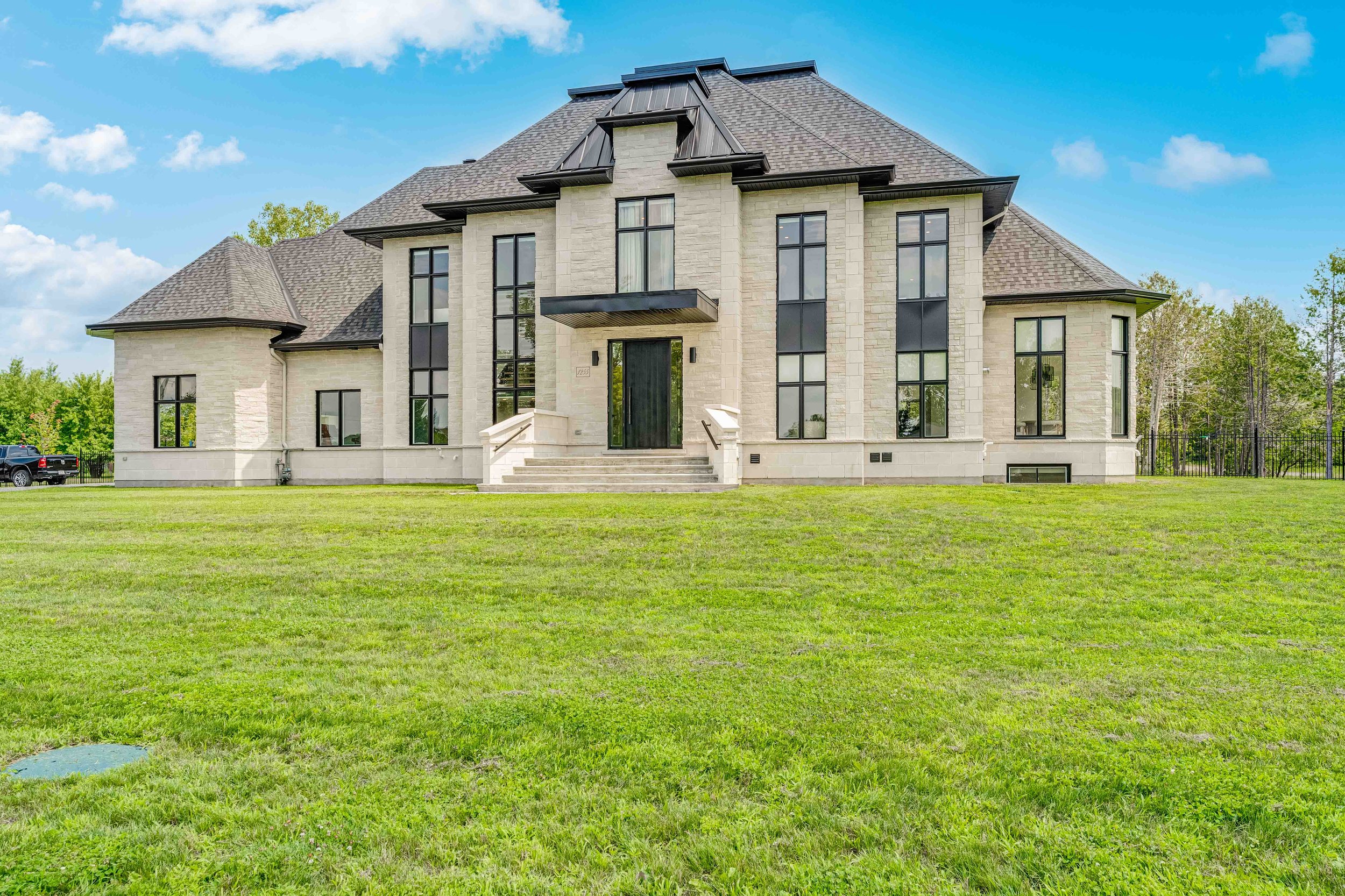 Large modern mansion with black-framed windows and light stone exterior, set against a blue sky, surrounded by a lush green lawn.