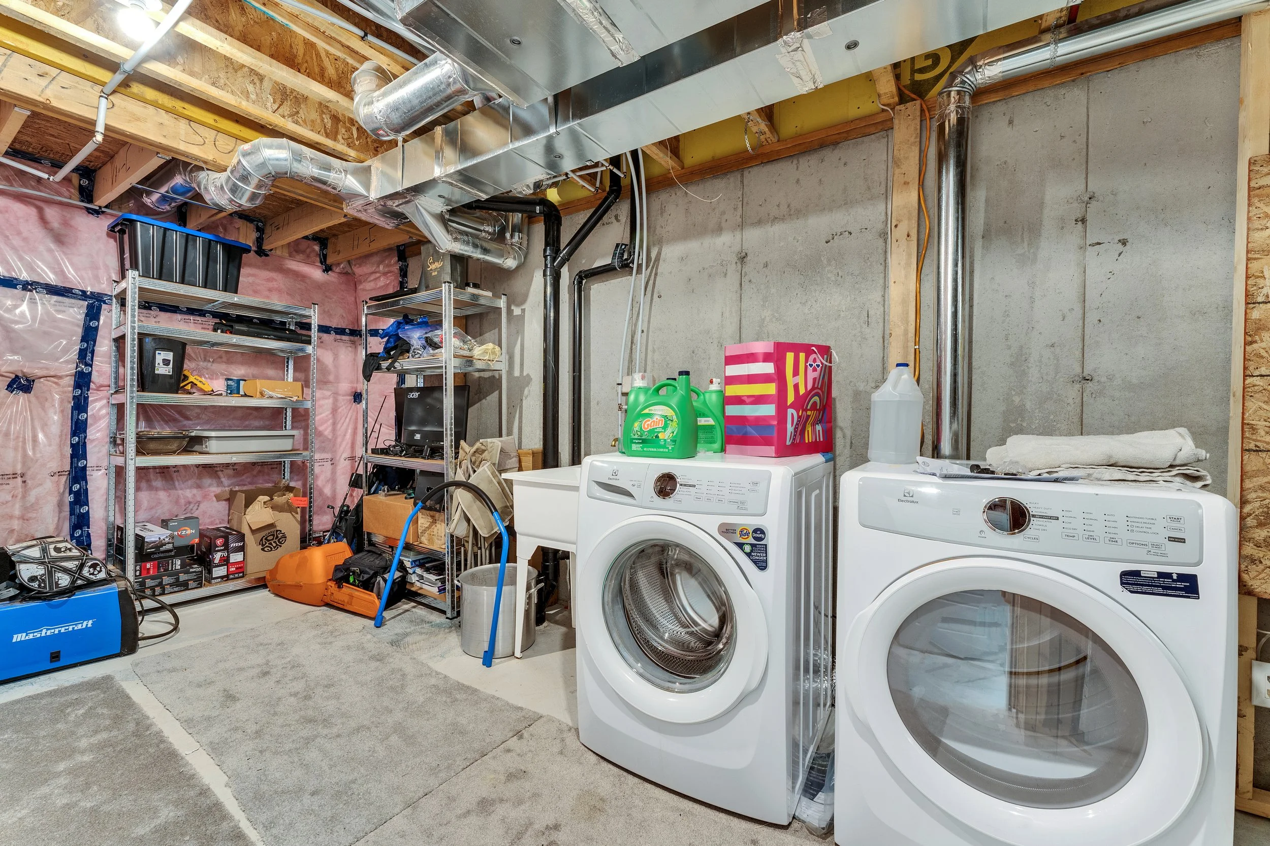 A basement laundry room with two front-loading washing machines, one with laundry supplies on top, and a metal shelving unit holding tools, boxes, and other items. There are exposed wooden framing, pipes, and ductwork on the ceiling, pink insulation 