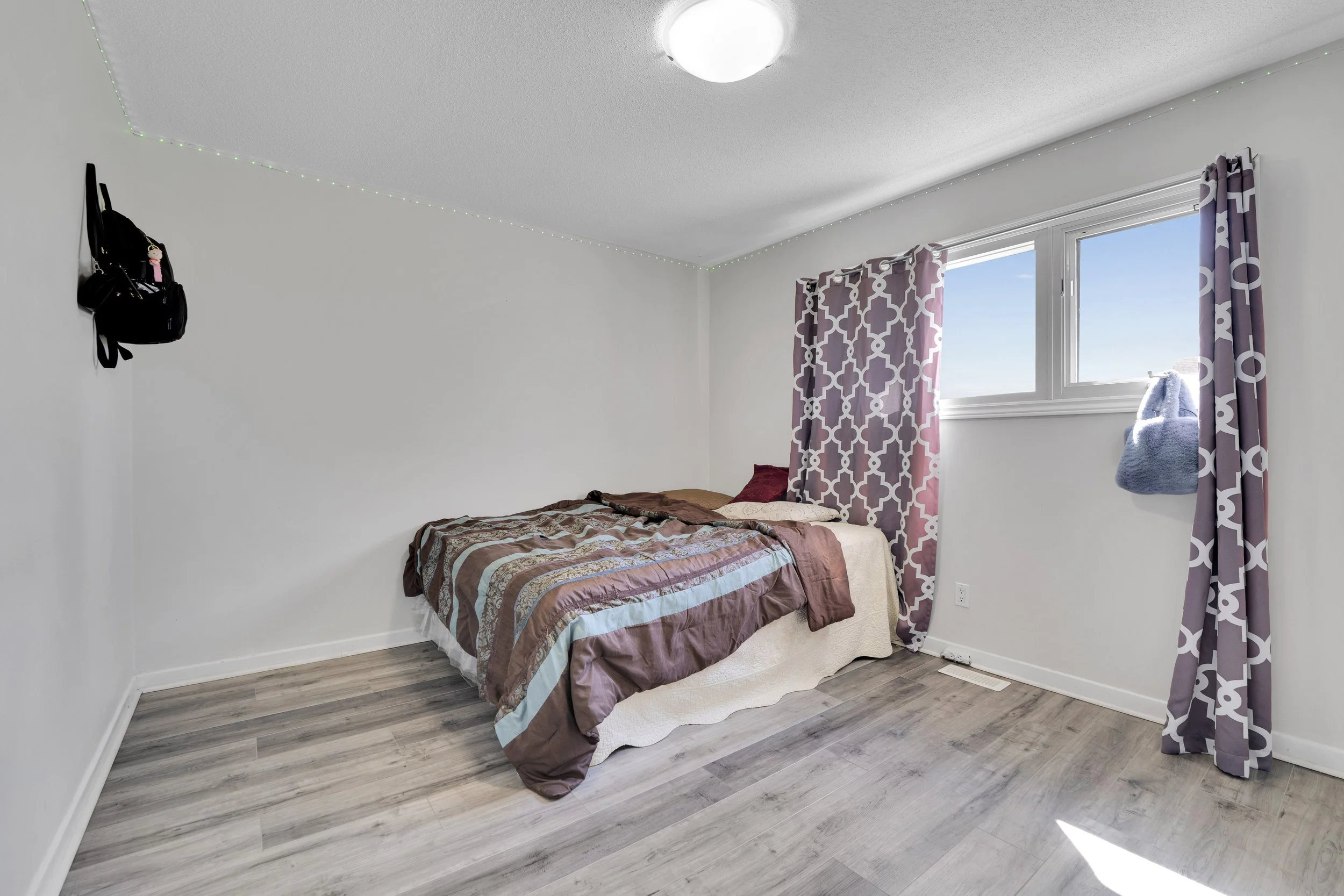 A bedroom with a bed that has a striped brown, pink, and blue blanket, two pillows, and a bed skirt. The window has pink and purple patterned curtains and a view of a clear sky outside. The floor is gray wood, and there is a black bag hanging on the 
