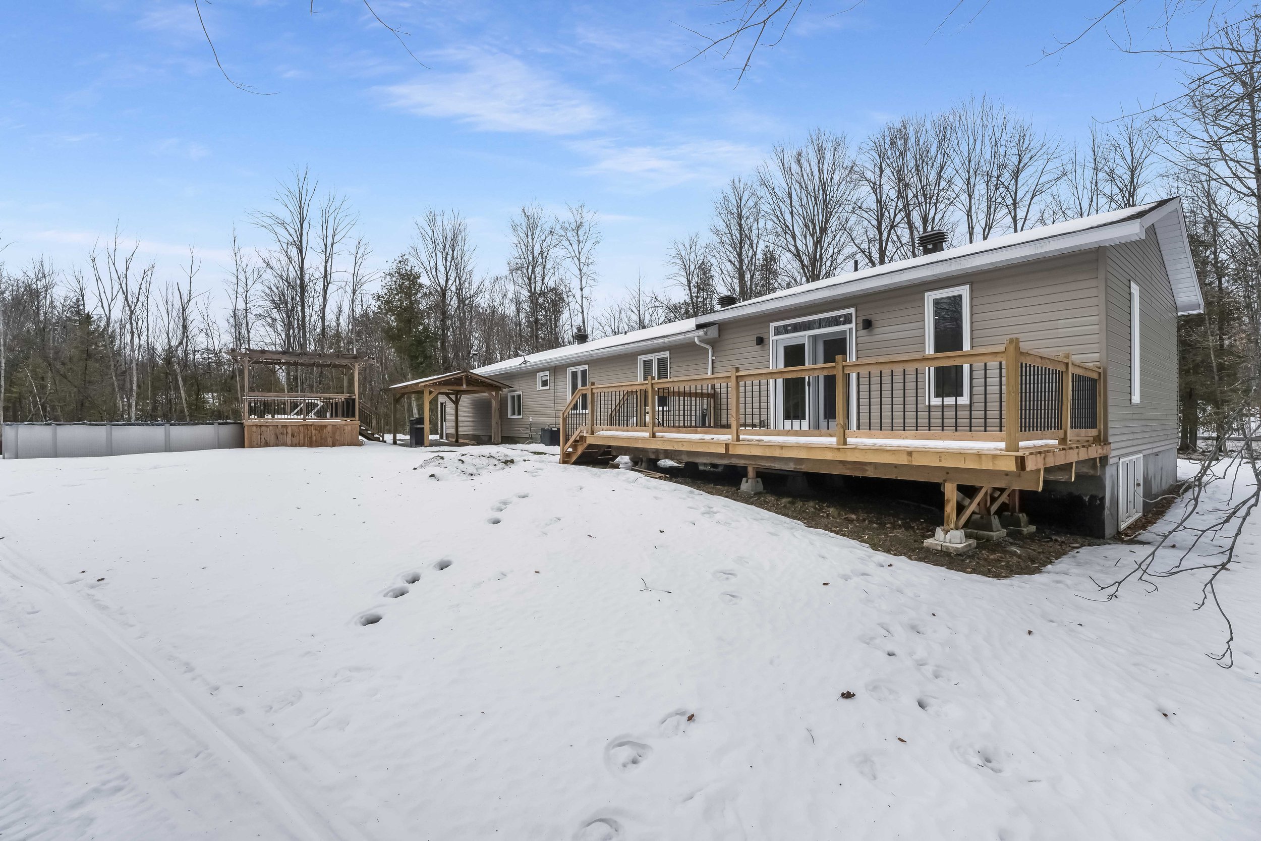 Backyard of a beige house with a wooden deck in winter, snow on the ground, leafless trees in the background, and clear blue sky. Real Estate Photography in Ottawa