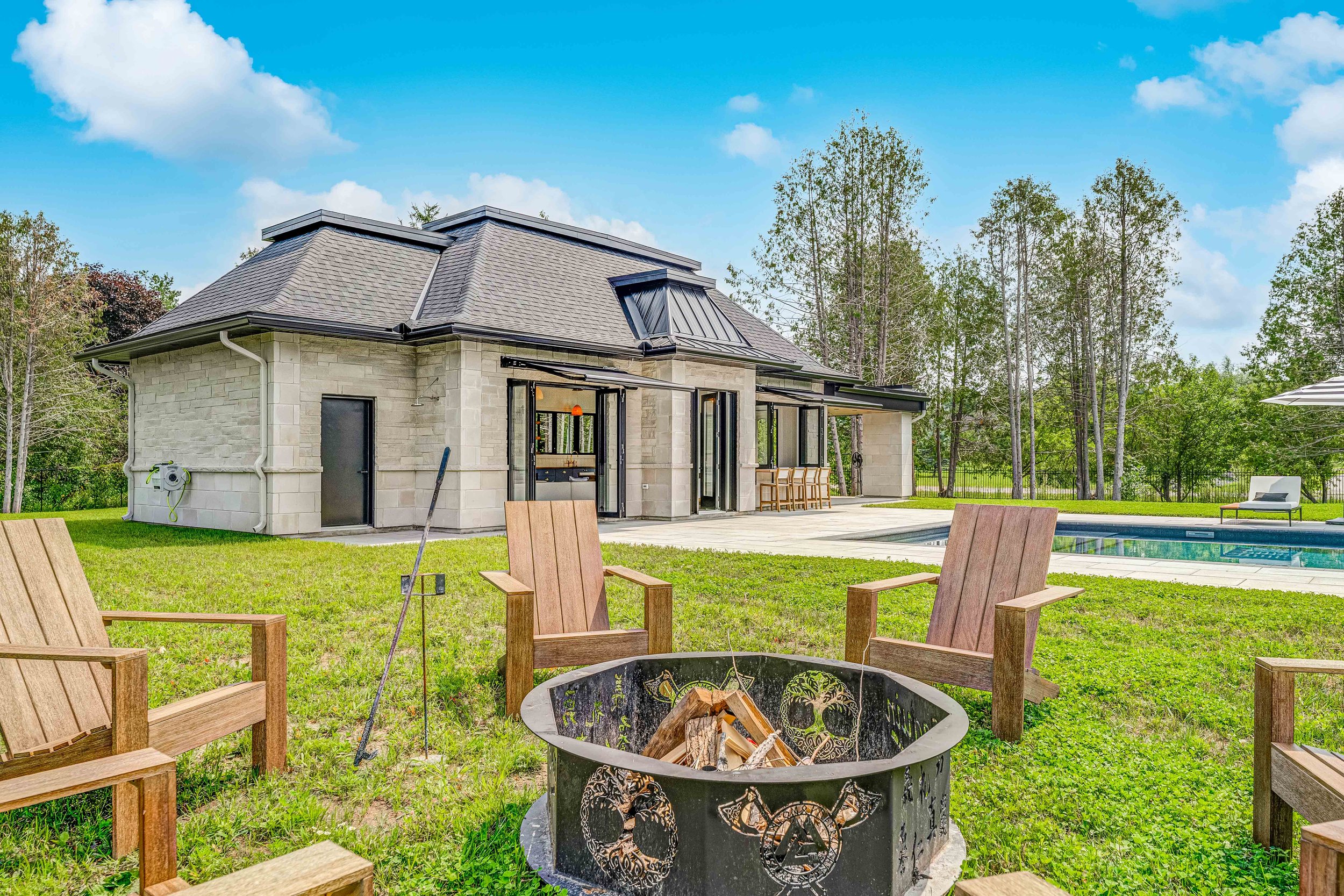 Modern house with a light stone exterior, surrounded by a grassy yard. In the foreground, there is a circular fire pit with wooden chairs around it. A swimming pool is visible to the right, accompanied by a lounge chair and an outdoor dining area und